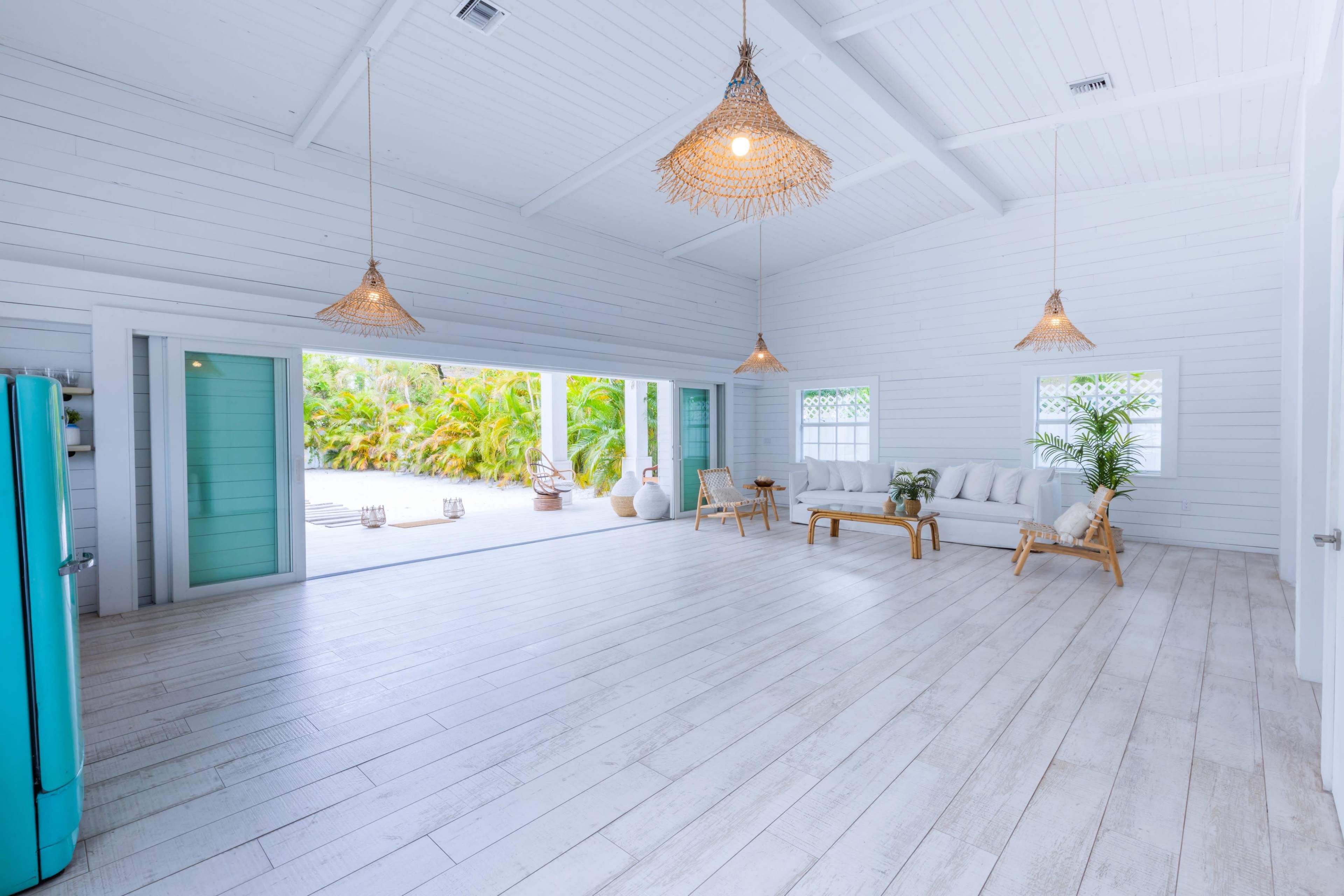 A spacious, brightly lit interior features white wooden walls, a seating area with a bamboo sofa, and tropical plants visible through large windows.
