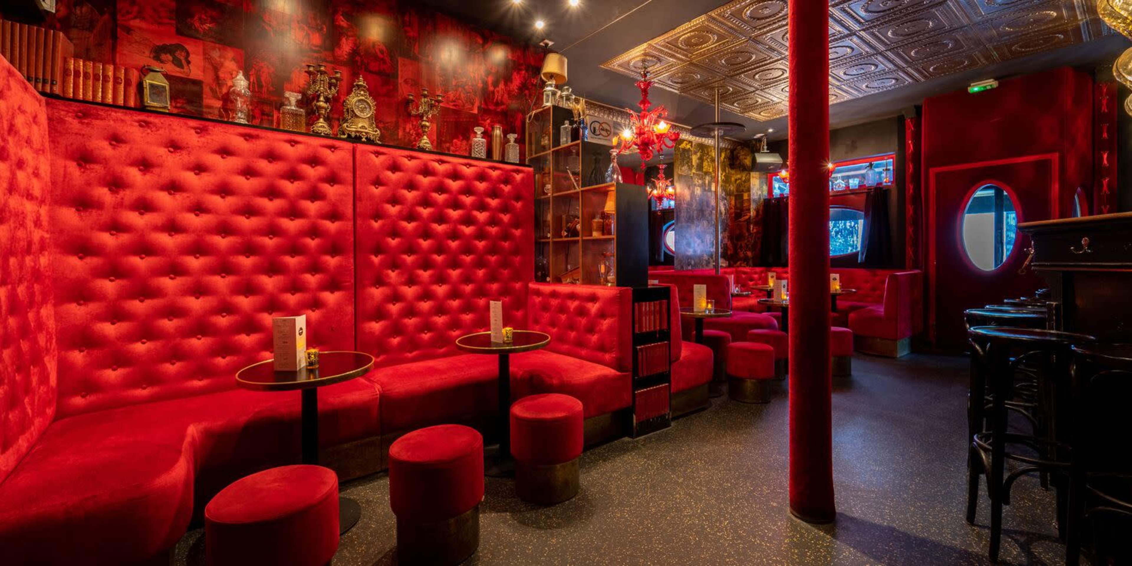 An interior of a stylish lounge featuring deep red tufted seating, small round tables, and decorative shelves with various items.