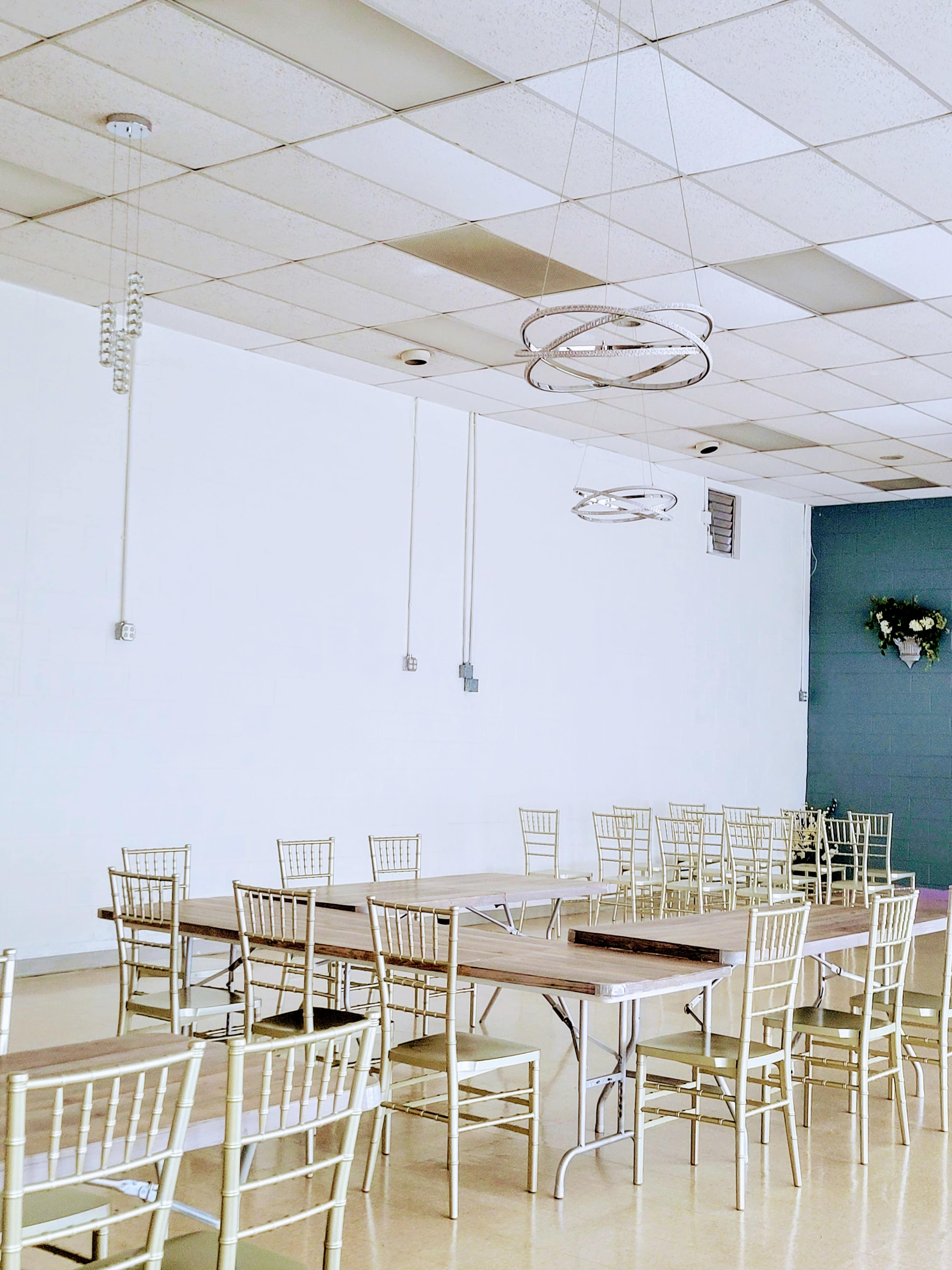 A spacious indoor venue features a long wooden table surrounded by gold chiavari chairs and pendant lighting.