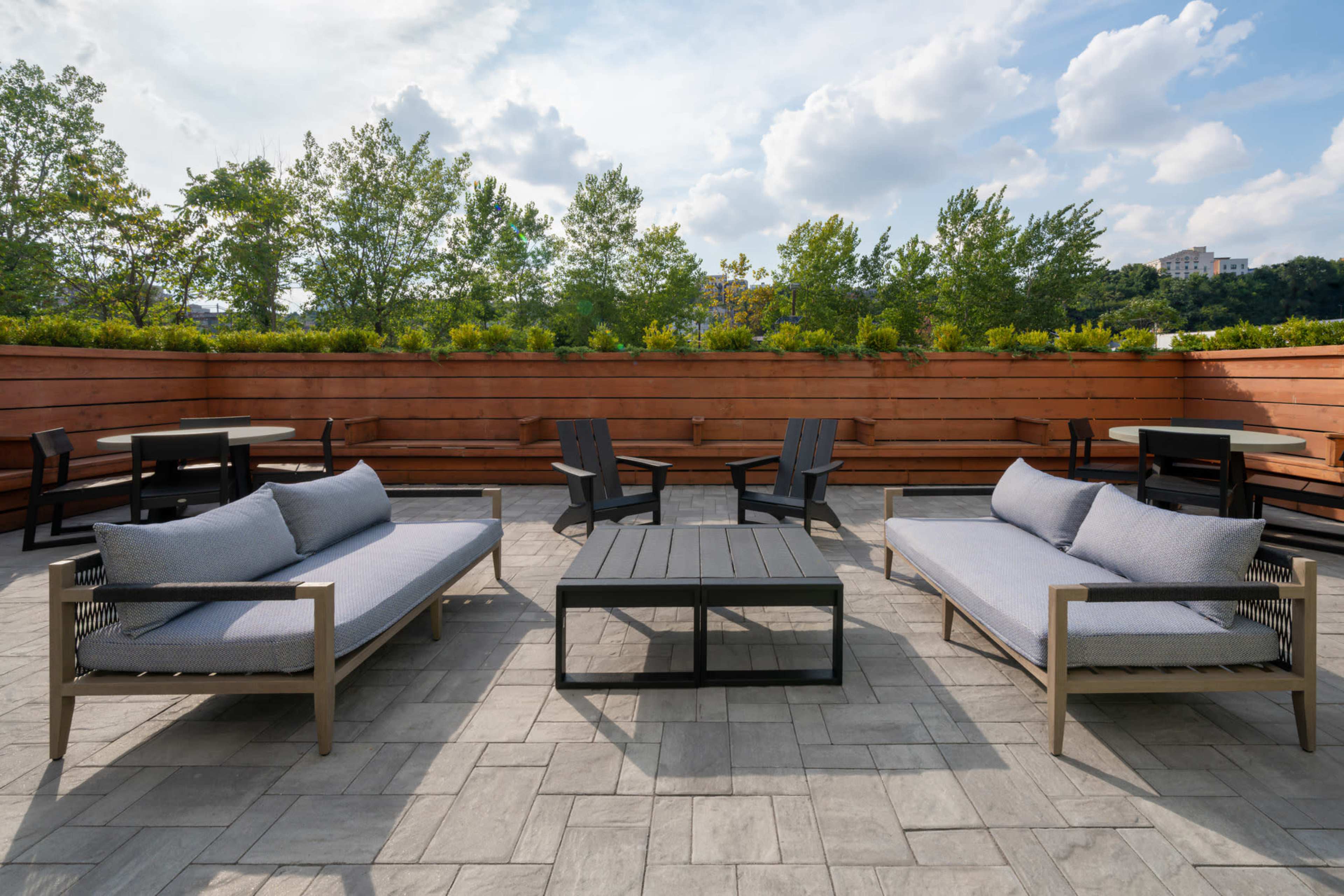 The image shows a rooftop patio featuring gray outdoor sofas and a black coffee table surrounded by green trees and partly cloudy skies.