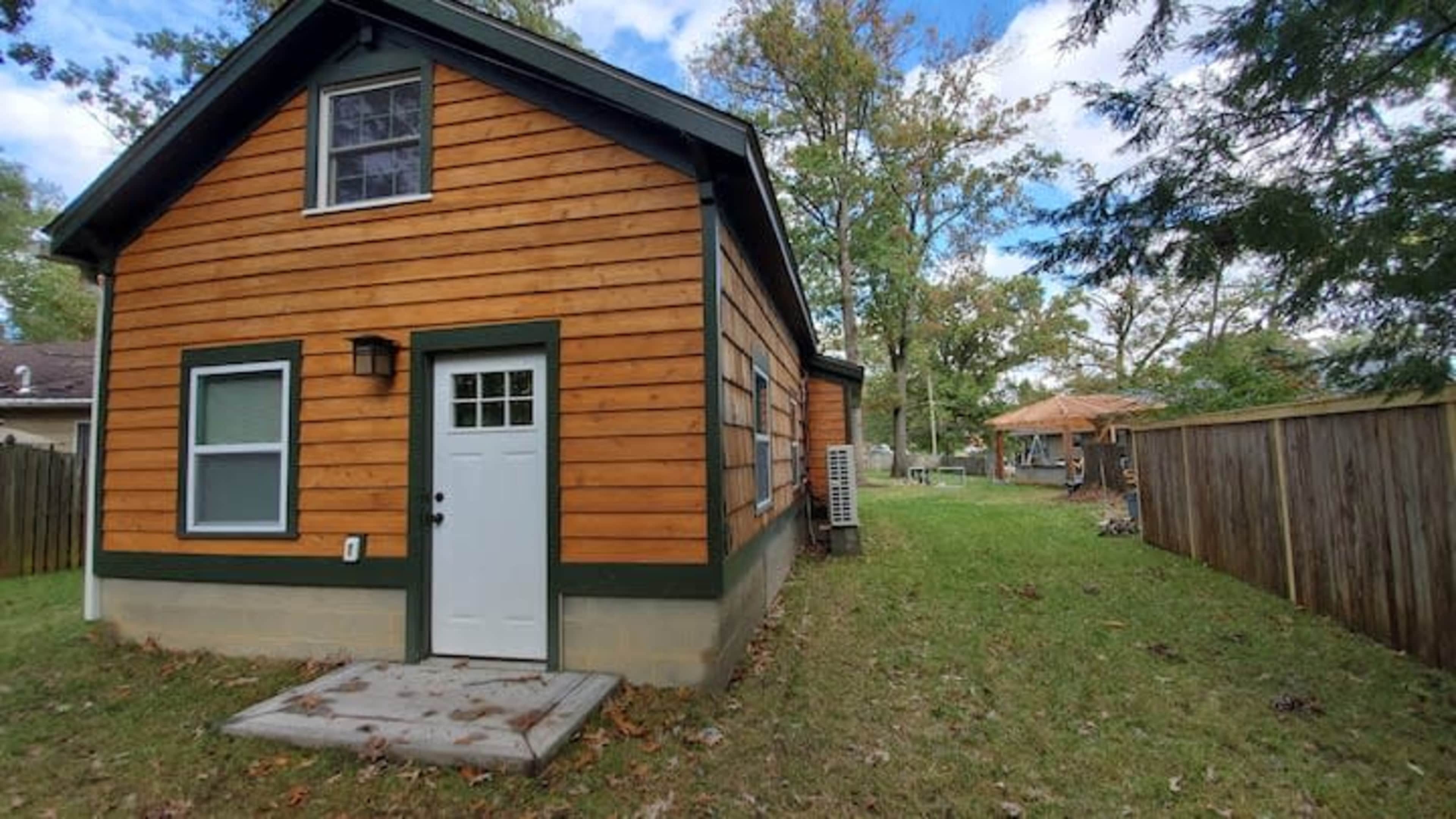 The image shows a small, two-story wooden house with a brown and green exterior, situated next to a grassy yard and a wooden fence.