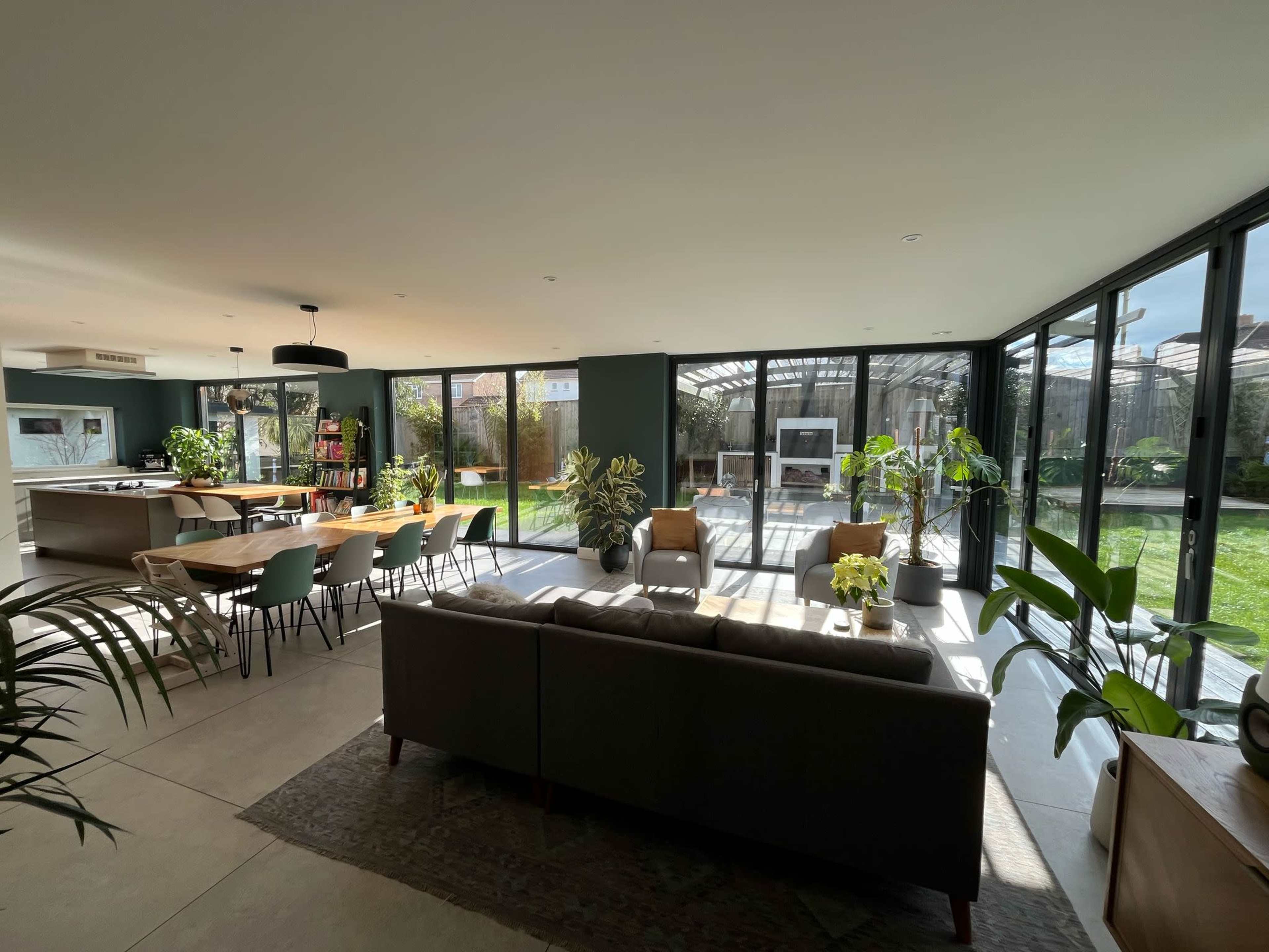 A modern open-concept living space features a dining area with a long table, a cozy sofa, and large glass walls that overlook a garden.