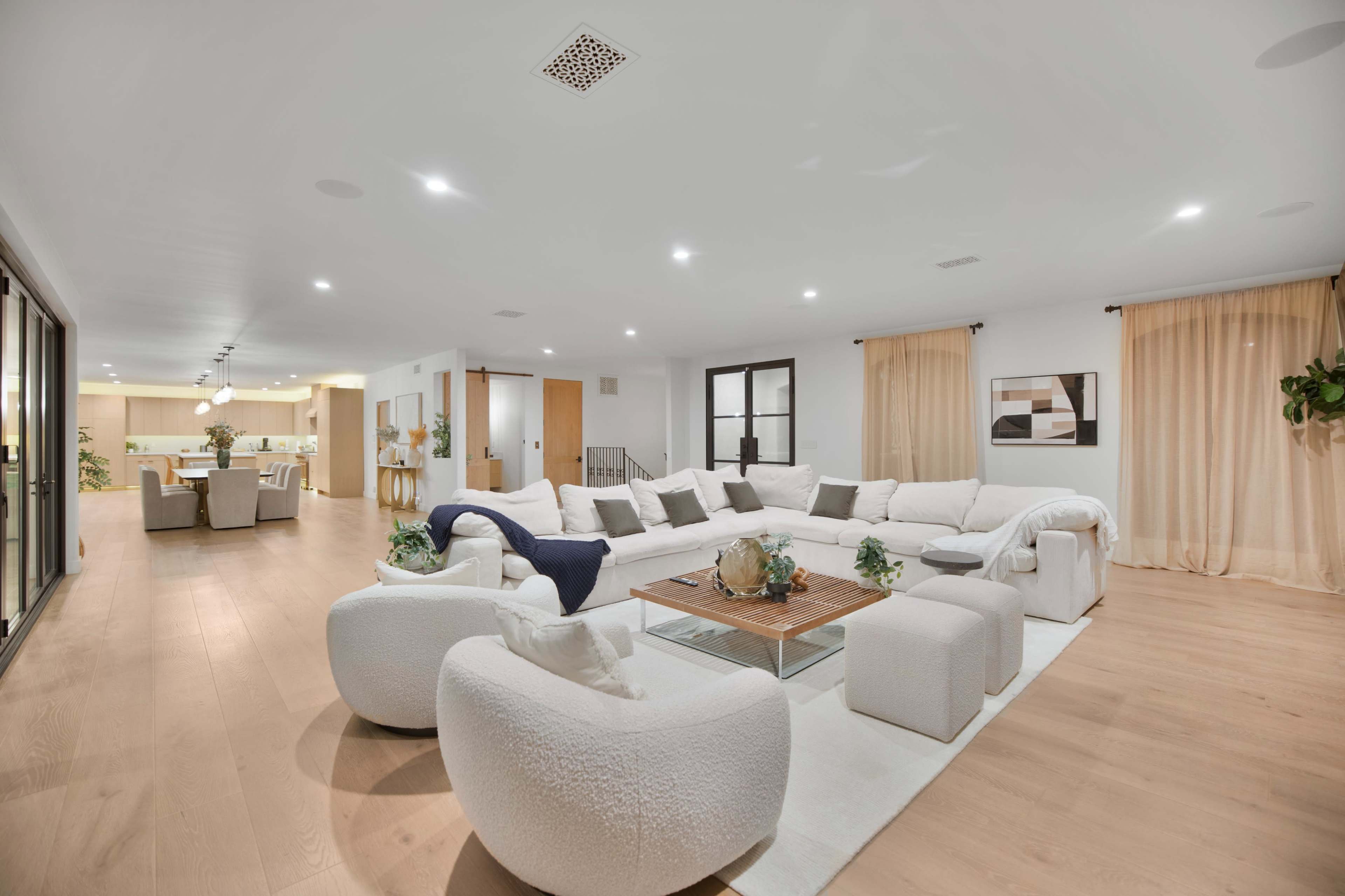 The image shows a modern, spacious living room furnished with a large, light-colored sectional sofa and a glass coffee table, leading to an open dining area visible in the background.