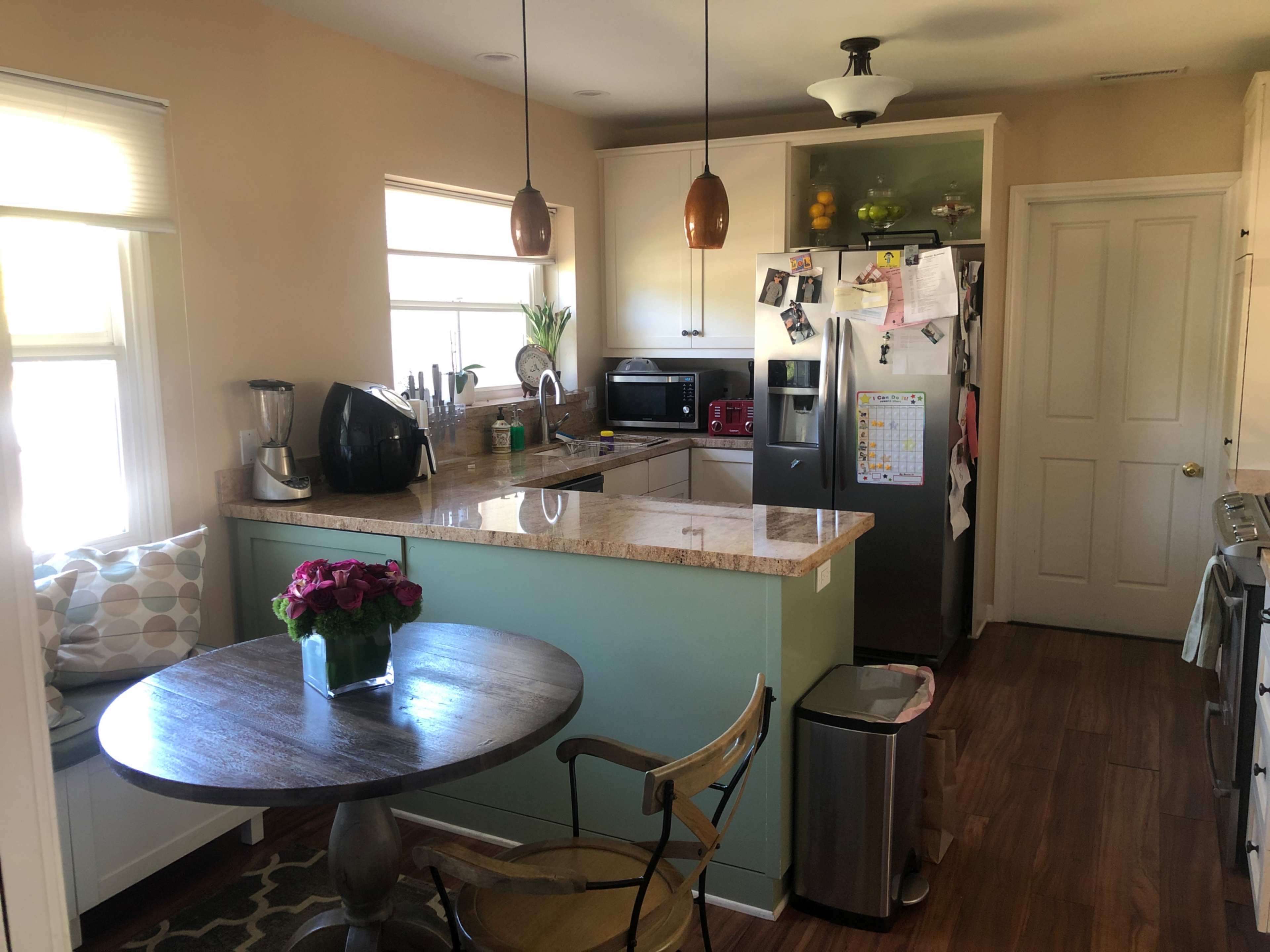 A modern kitchen features light-colored cabinets, a granite countertop, a table with flowers, and appliances including a refrigerator and microwave.