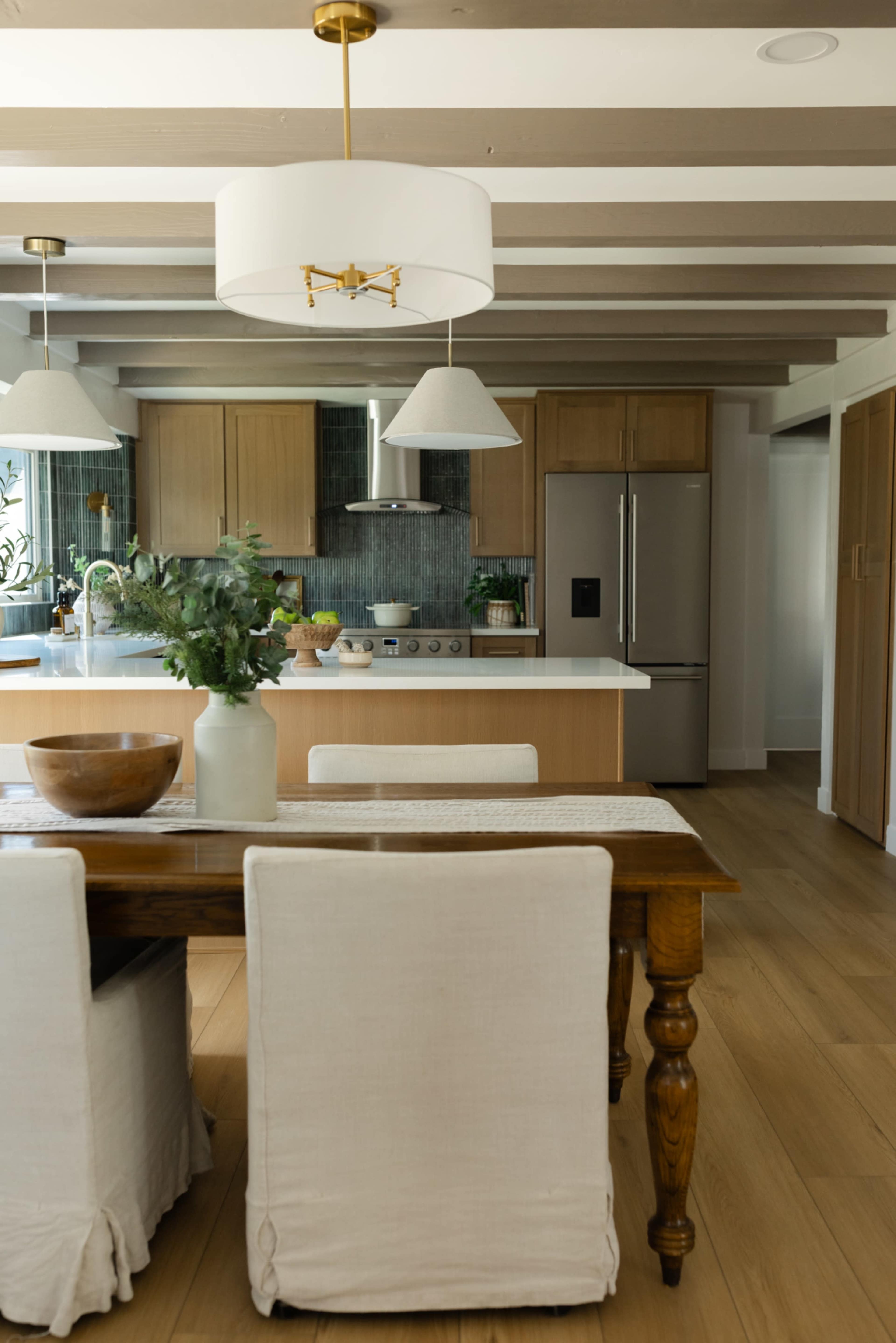 A modern kitchen features a wooden dining table with white chairs, stainless steel appliances, and an island topped with a bowl of greenery.