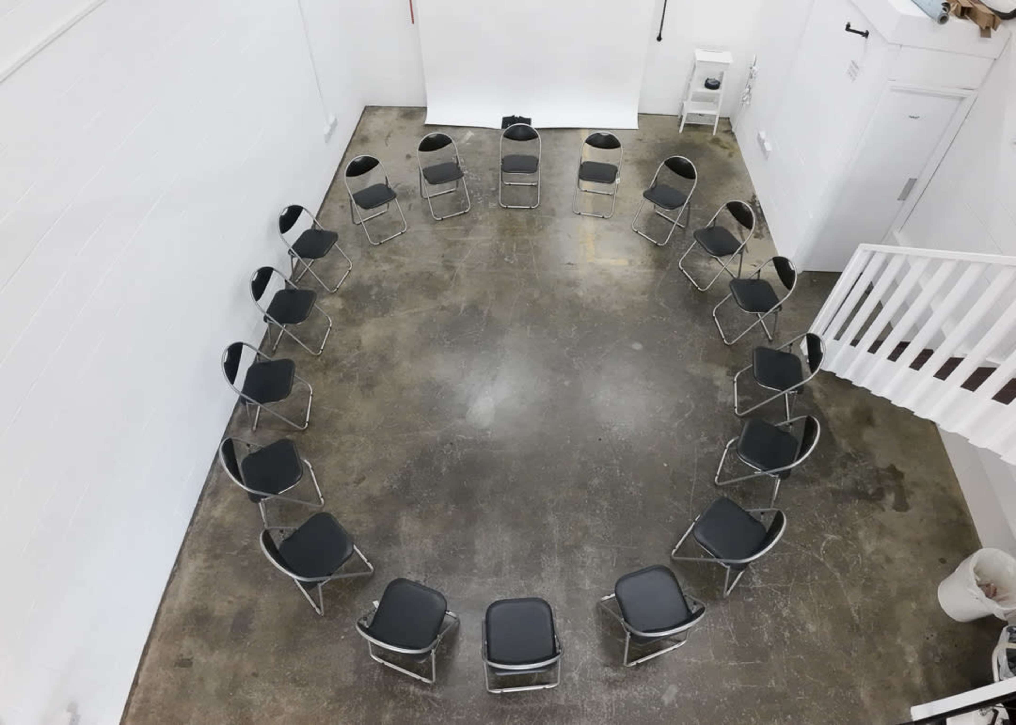 A circular arrangement of black chairs on a concrete floor inside a white-walled room.