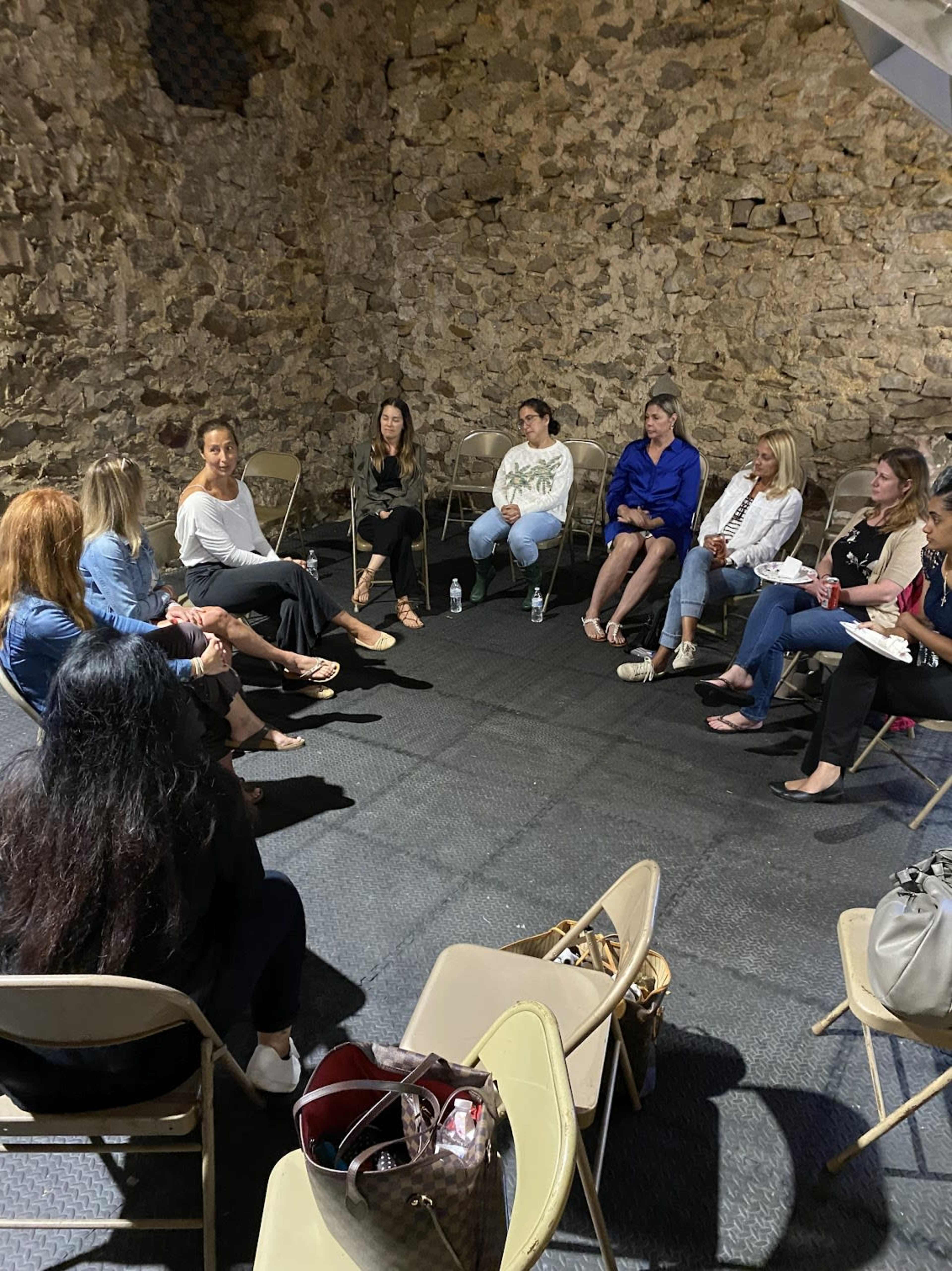 A group of women sits in a circle on folding chairs in a room with stone walls.