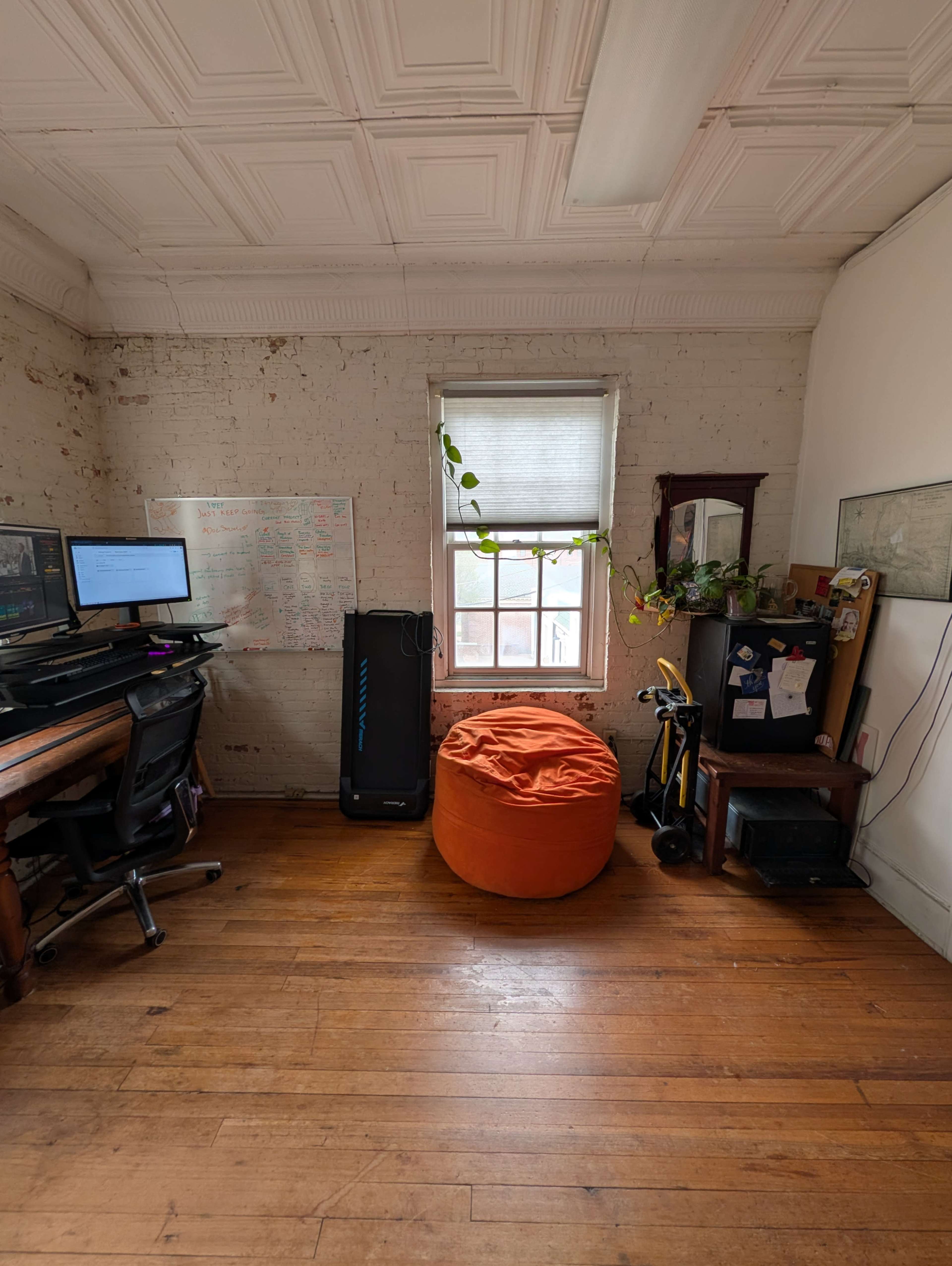 The image shows a small office with wooden floors, a desk with two computer monitors, a bean bag chair, and a window with a plant hanging near it.