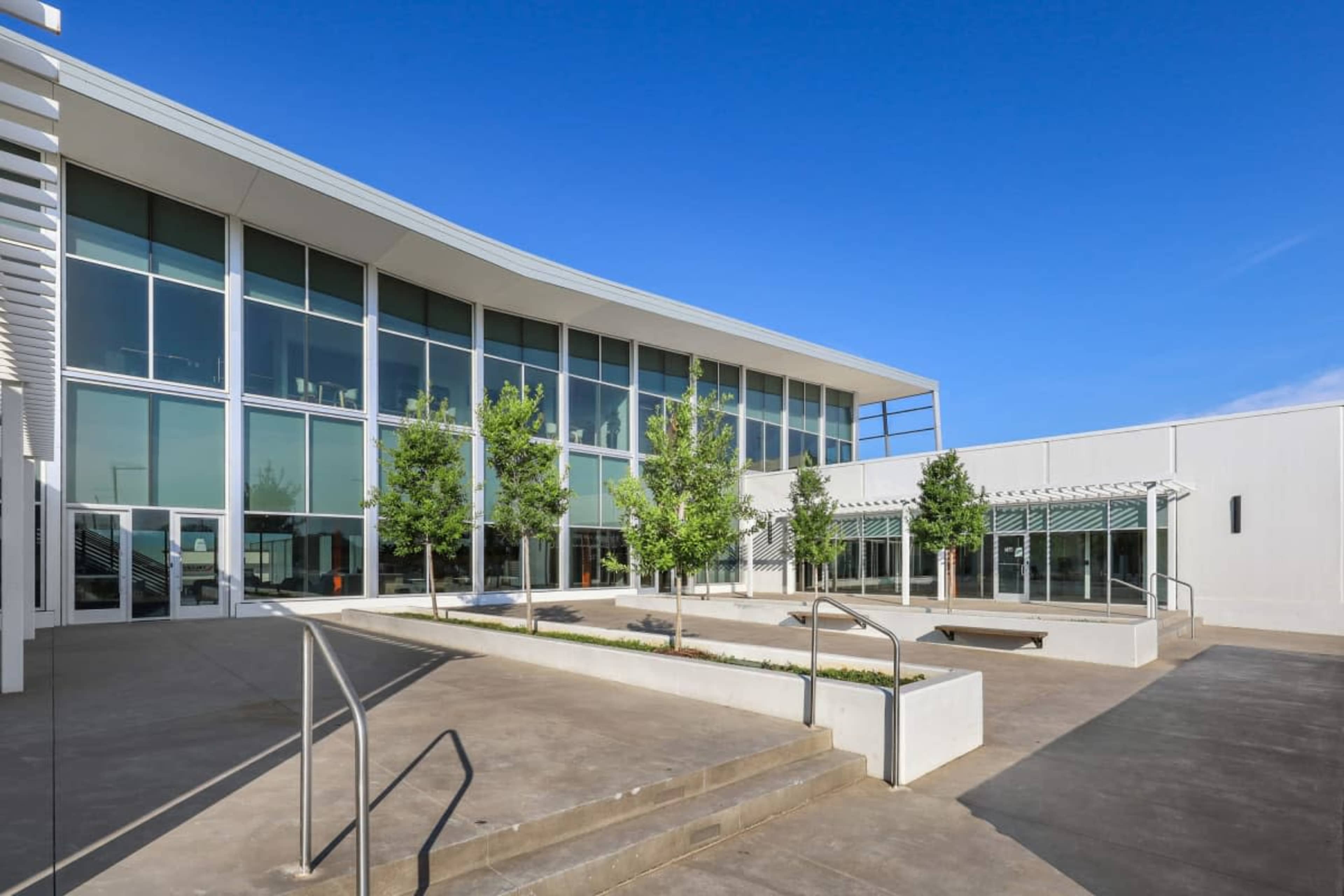 The image shows a modern building with large glass windows, surrounded by landscaped areas with small trees and concrete walkways.