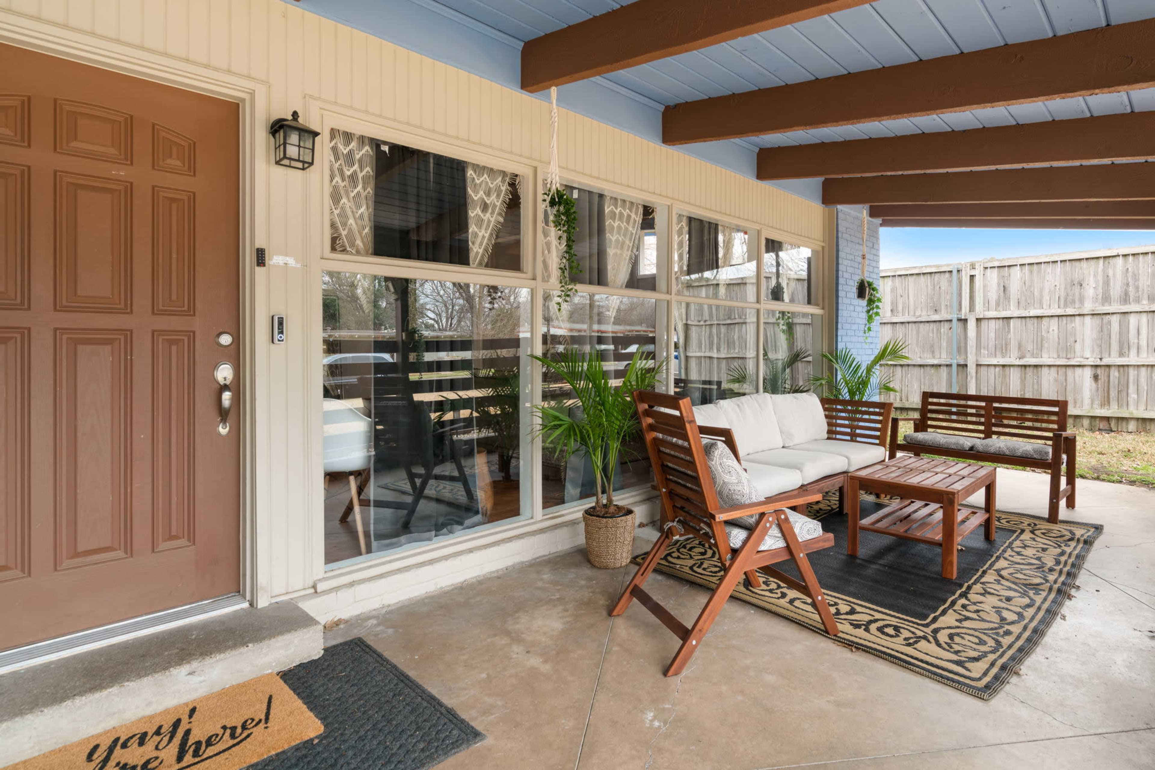 A covered porch with a wooden seating arrangement, a coffee table, and large windows overlooking a backyard enclosed by a wooden fence.