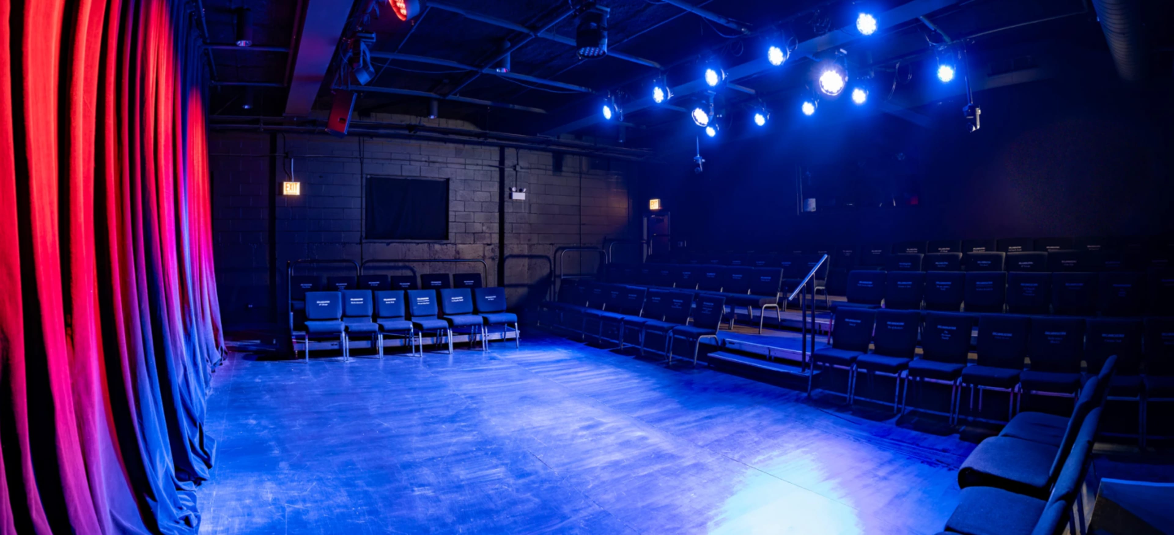 The image shows a dimly lit theater stage with a curtain to the left and rows of empty seats arranged in front.