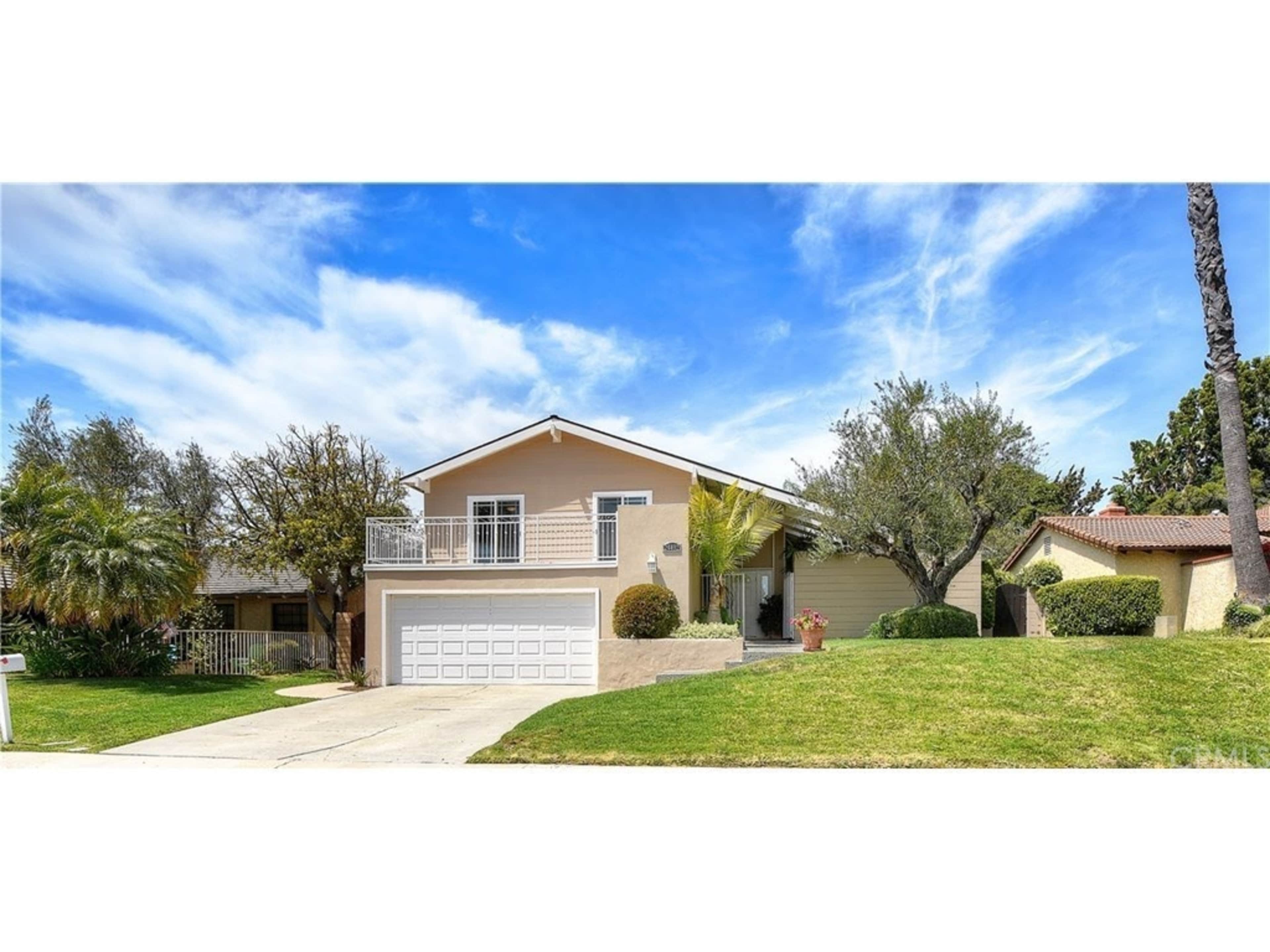 A single-story house with a white garage door and a balcony is set amidst green lawns and palm trees under a blue sky.
