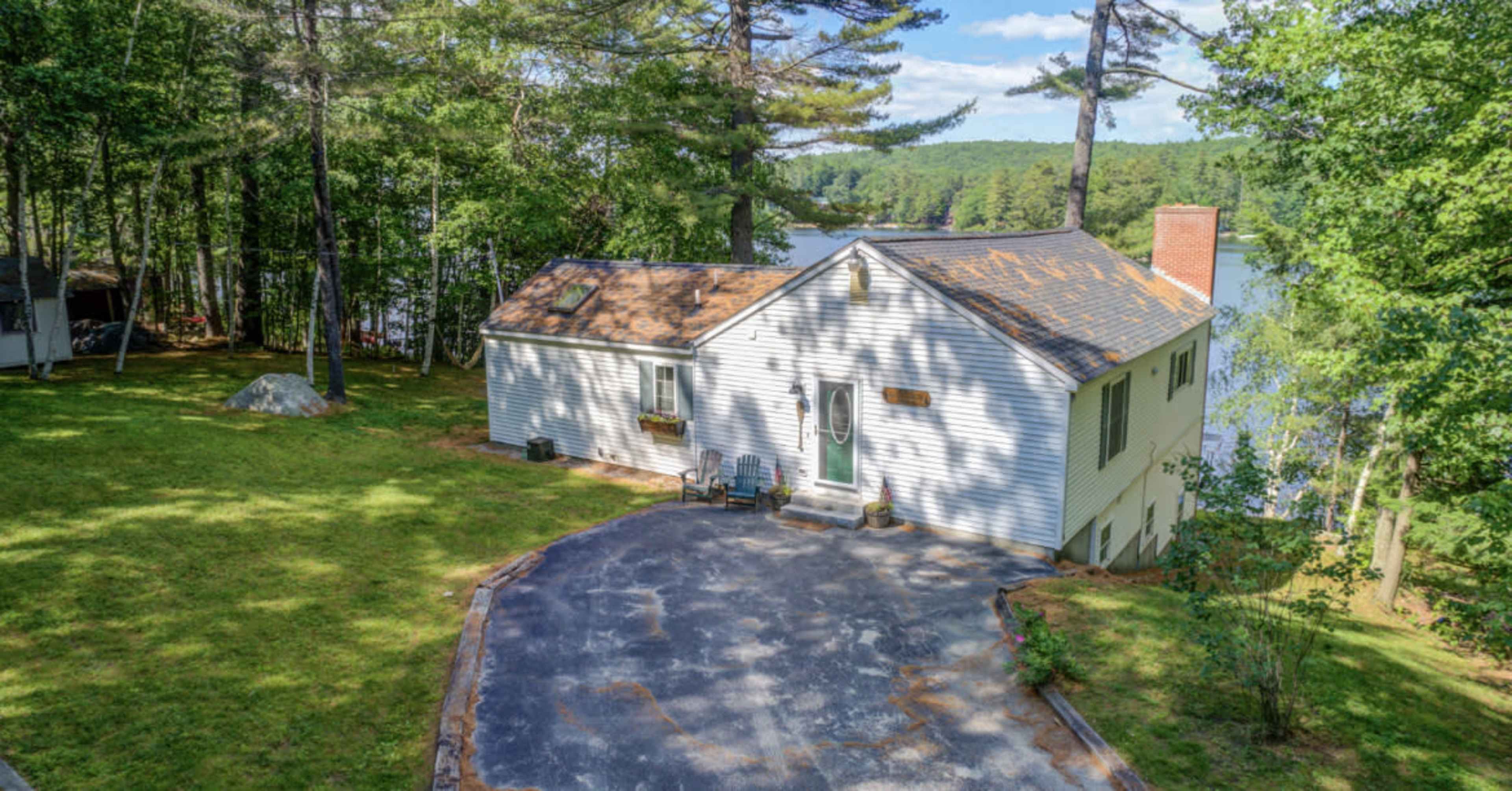 A single-story house with a gravel driveway is situated near a lake, surrounded by trees.