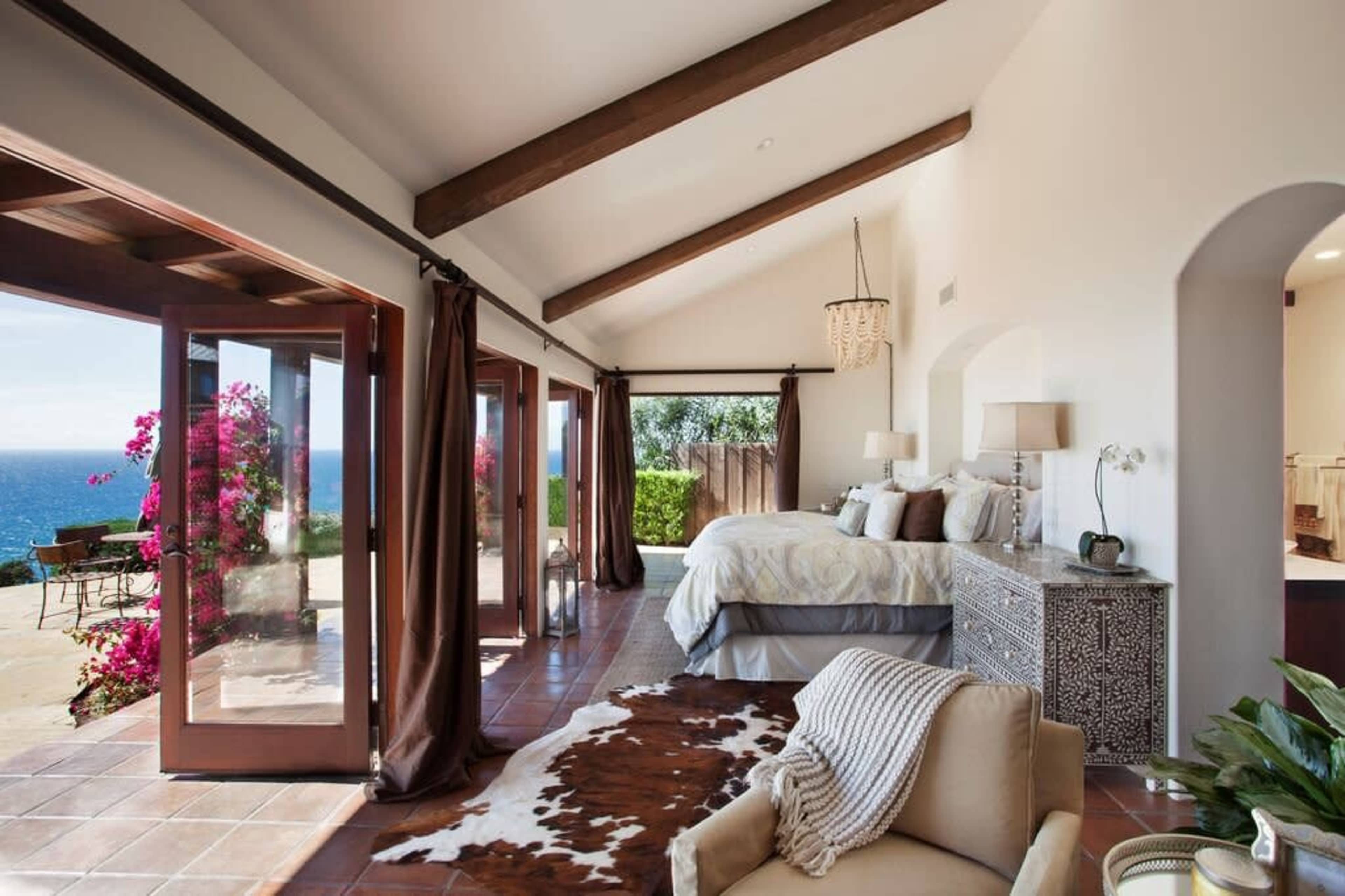 A spacious bedroom features a large bed, wooden beams on the ceiling, and sliding glass doors opening to a patio with ocean views and flowering plants.
