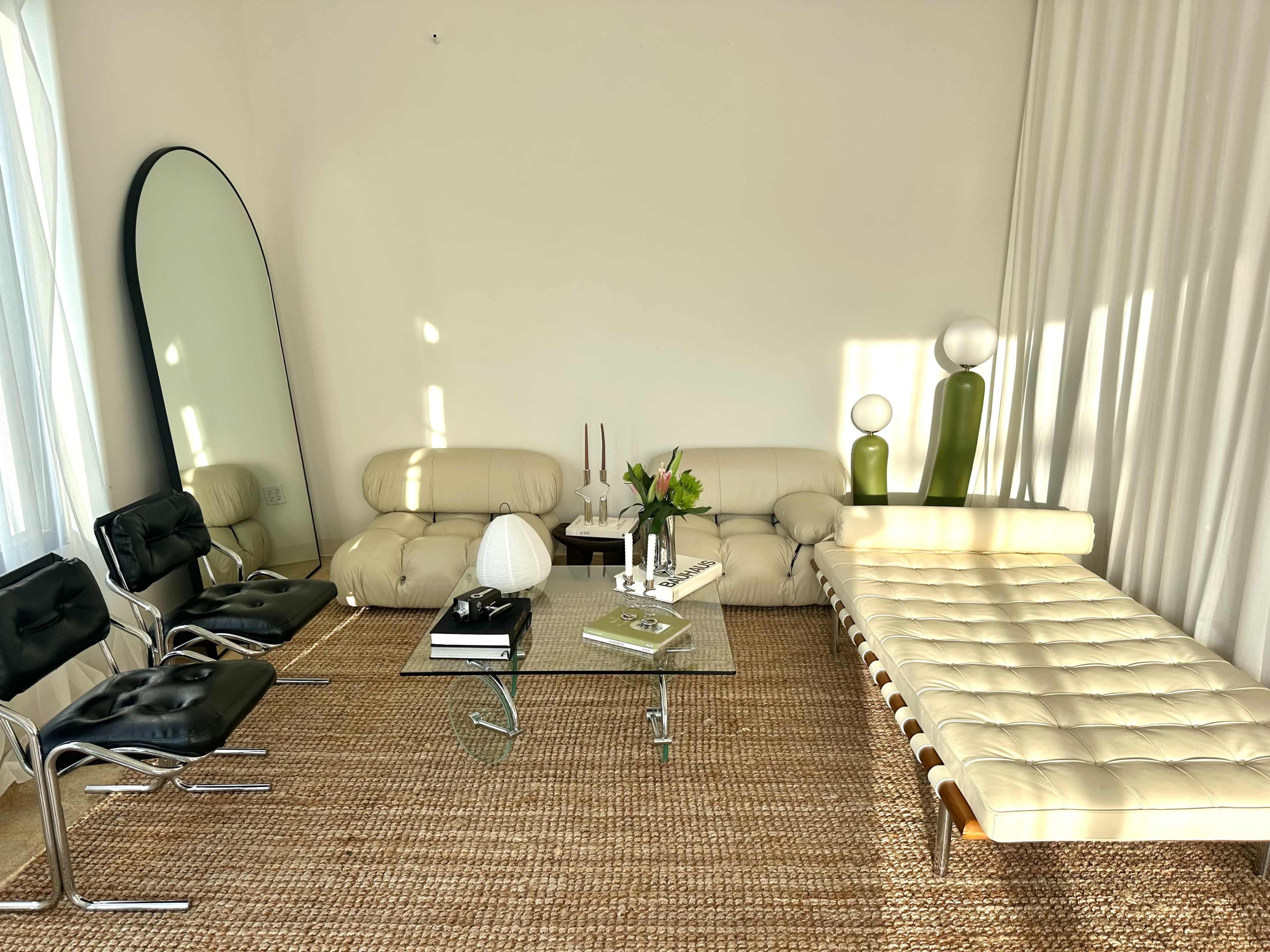 The room features a modern seating area with a cream-colored sofa, black chairs, a glass coffee table, and decorative items, all illuminated by natural light.