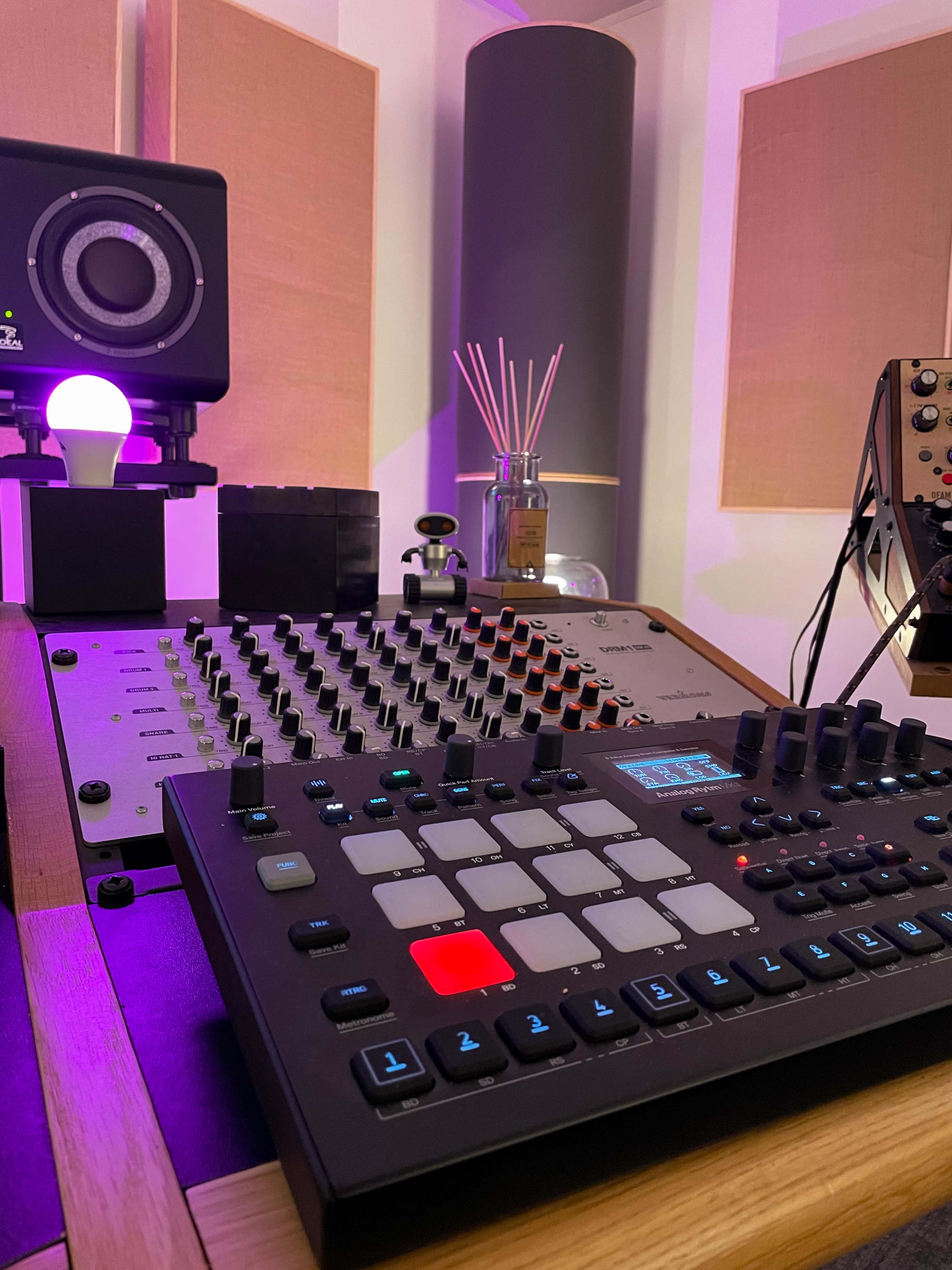 The image shows a close-up of a music production setup featuring a mixer, a drum machine, and ambient lighting in a studio environment.