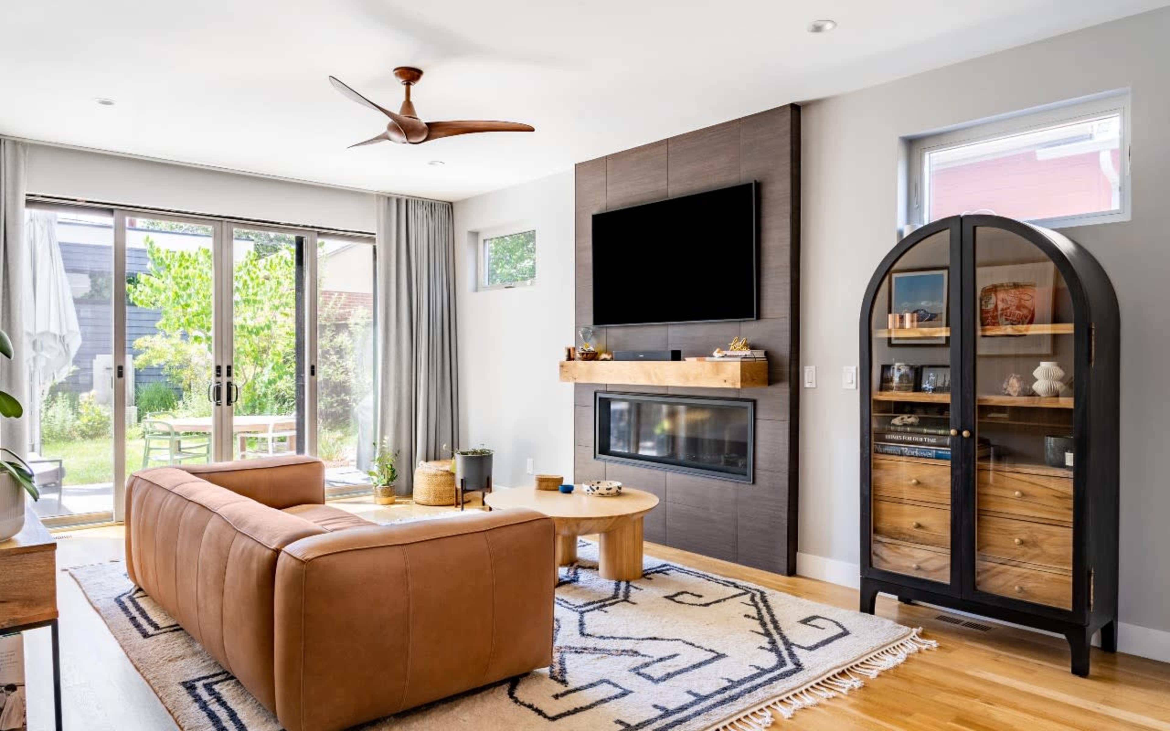 A modern living room features a brown leather sofa, a wall-mounted television above a sleek fireplace, and a glass-front cabinet displaying decorative items, with sliding doors leading to an outdoor space.