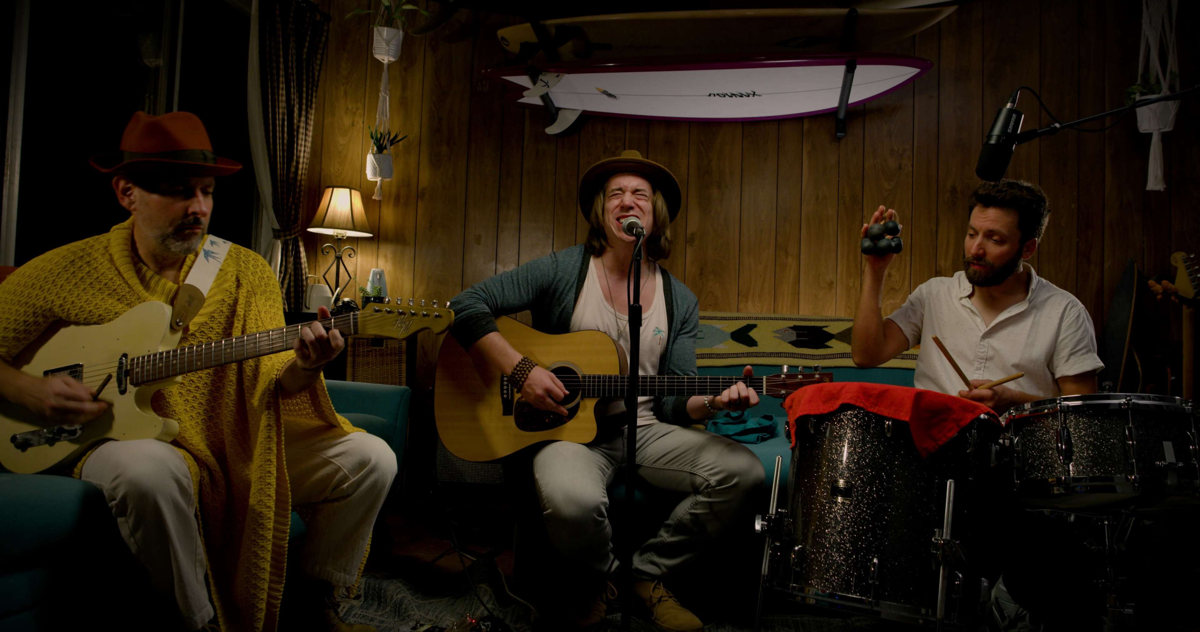 Three musicians perform in a cozy room, with one playing guitar, another singing, and the third playing drums.