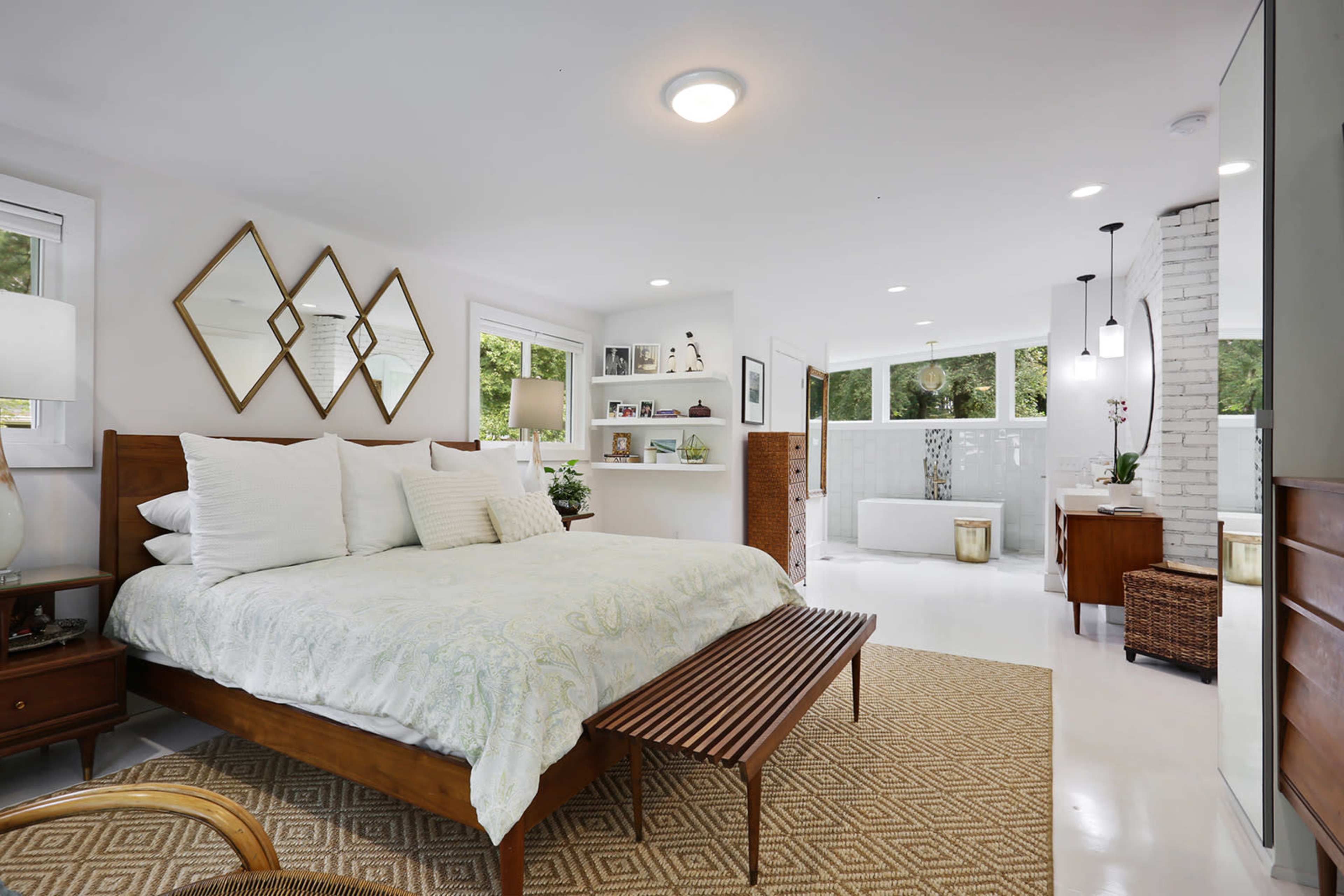 A modern bedroom featuring a large bed with decorative pillows, a wooden bench at the foot, and a spacious adjoining bathroom with a freestanding tub.
