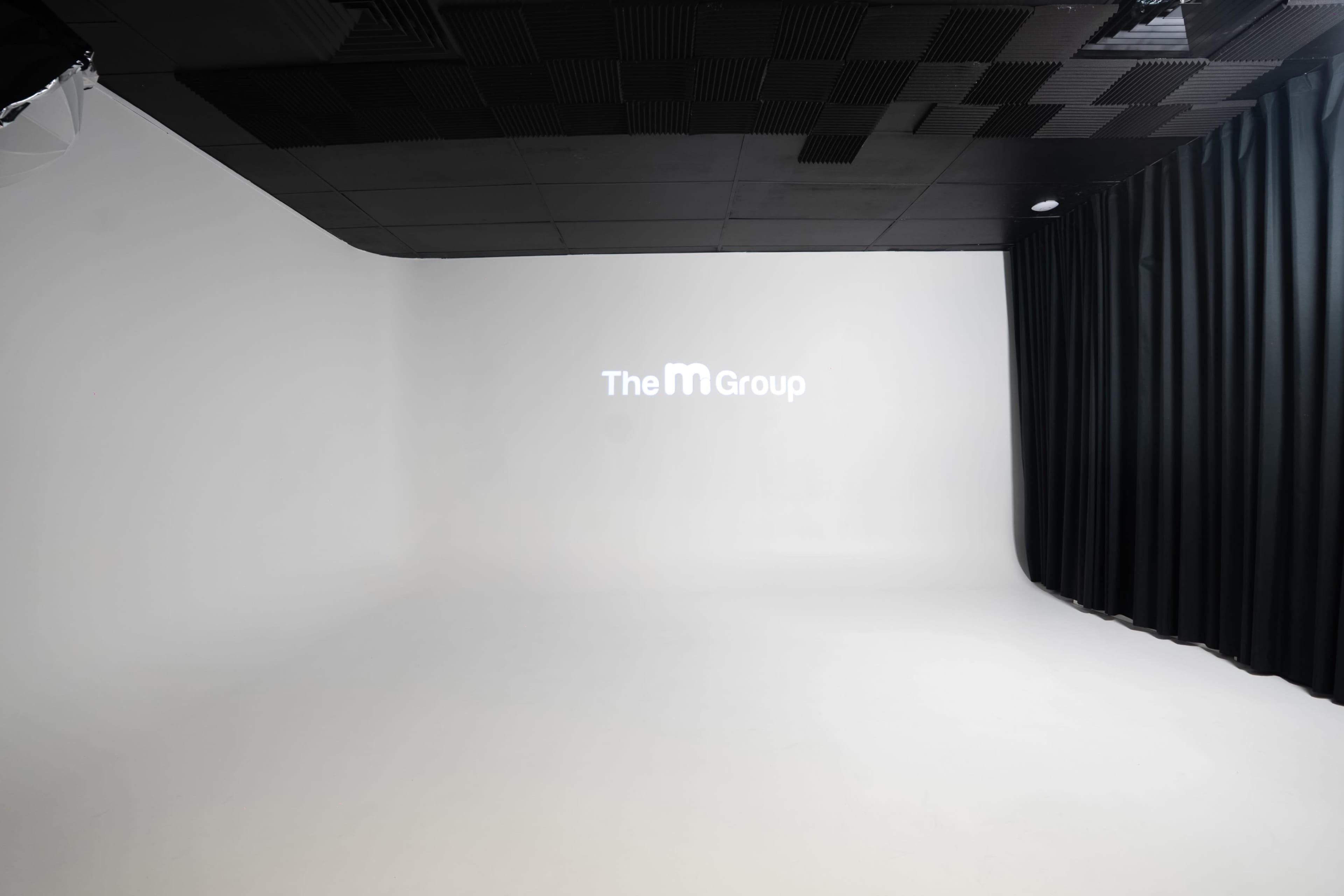 The image shows a minimalist studio space with a white backdrop, a black ceiling, and dark curtains on one side, featuring the text "The M Group" projected onto the wall.