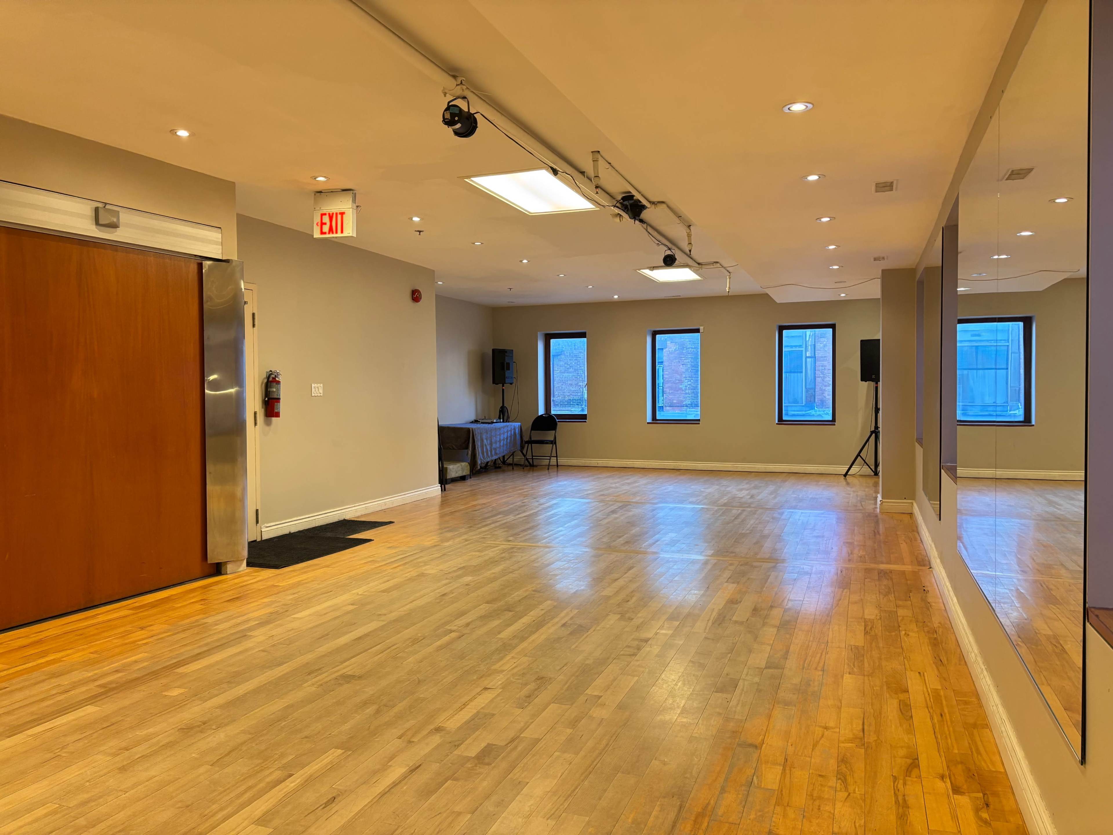 The image shows a spacious room with wooden flooring, equipped with windows, a couch, and sound equipment.