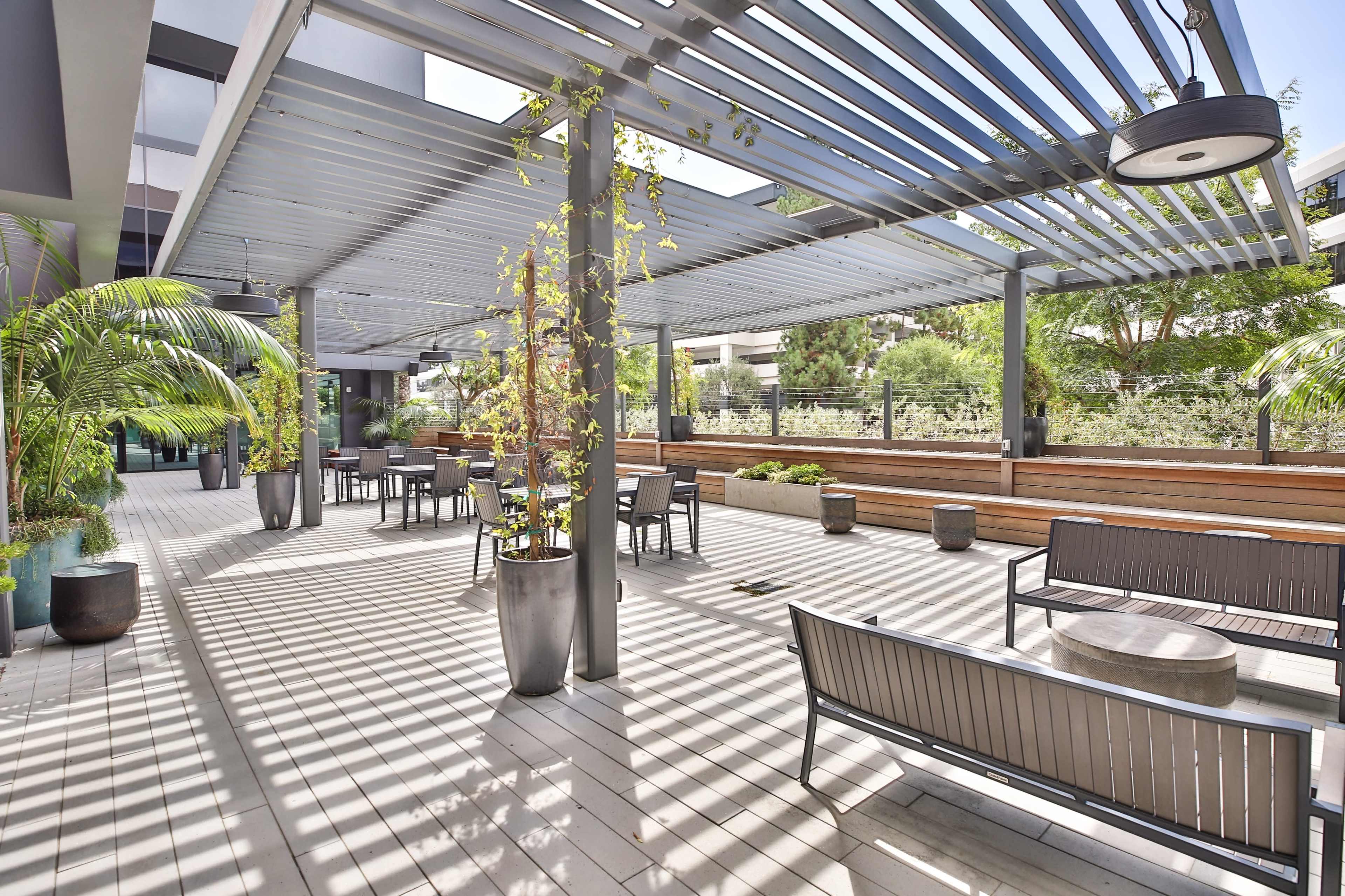 A modern outdoor patio area with benches, tables, and potted plants under a slatted pergola.