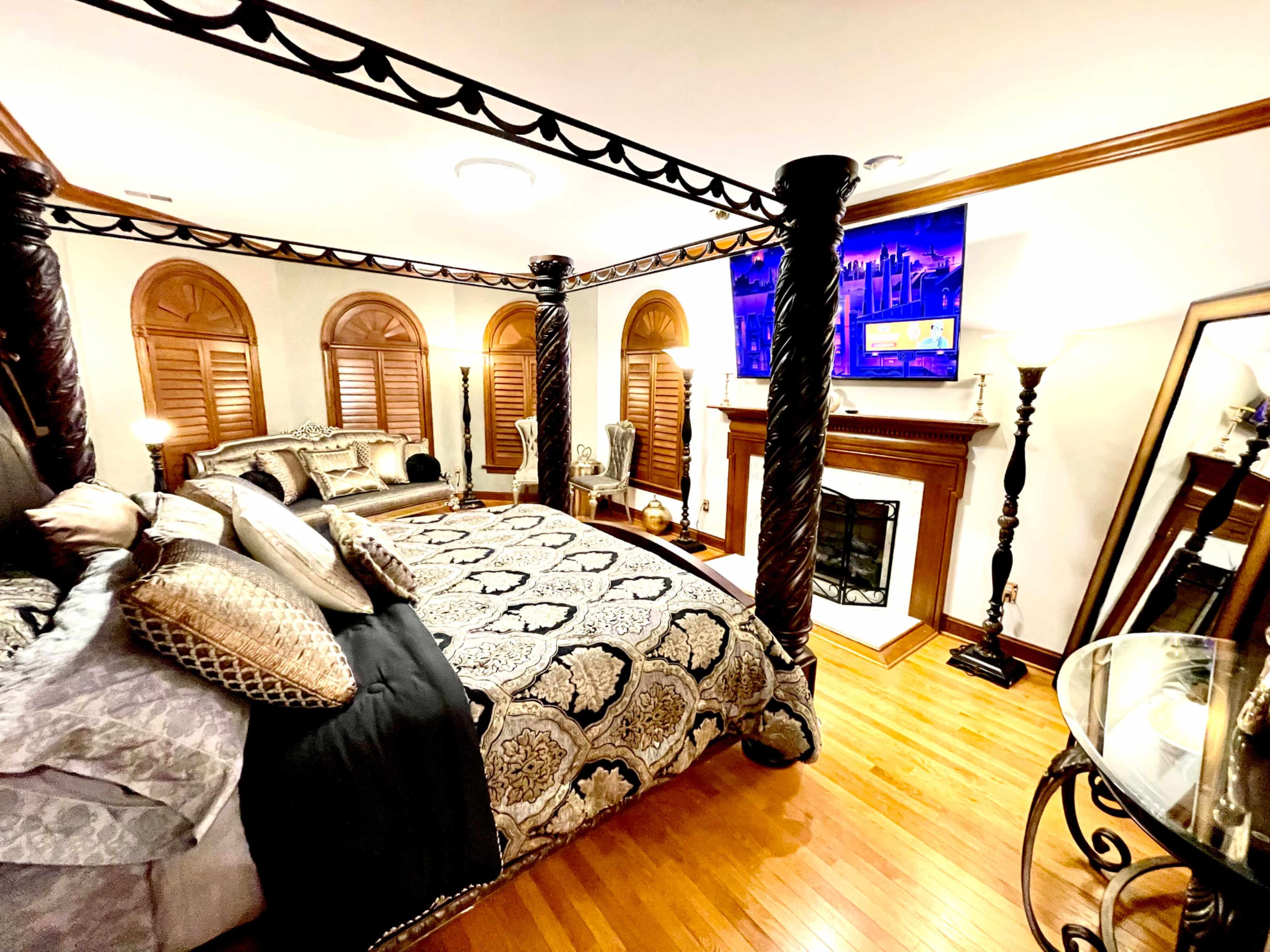 The image shows a well-furnished bedroom featuring a large four-poster bed, elegant decor, wooden floors, and a fireplace.