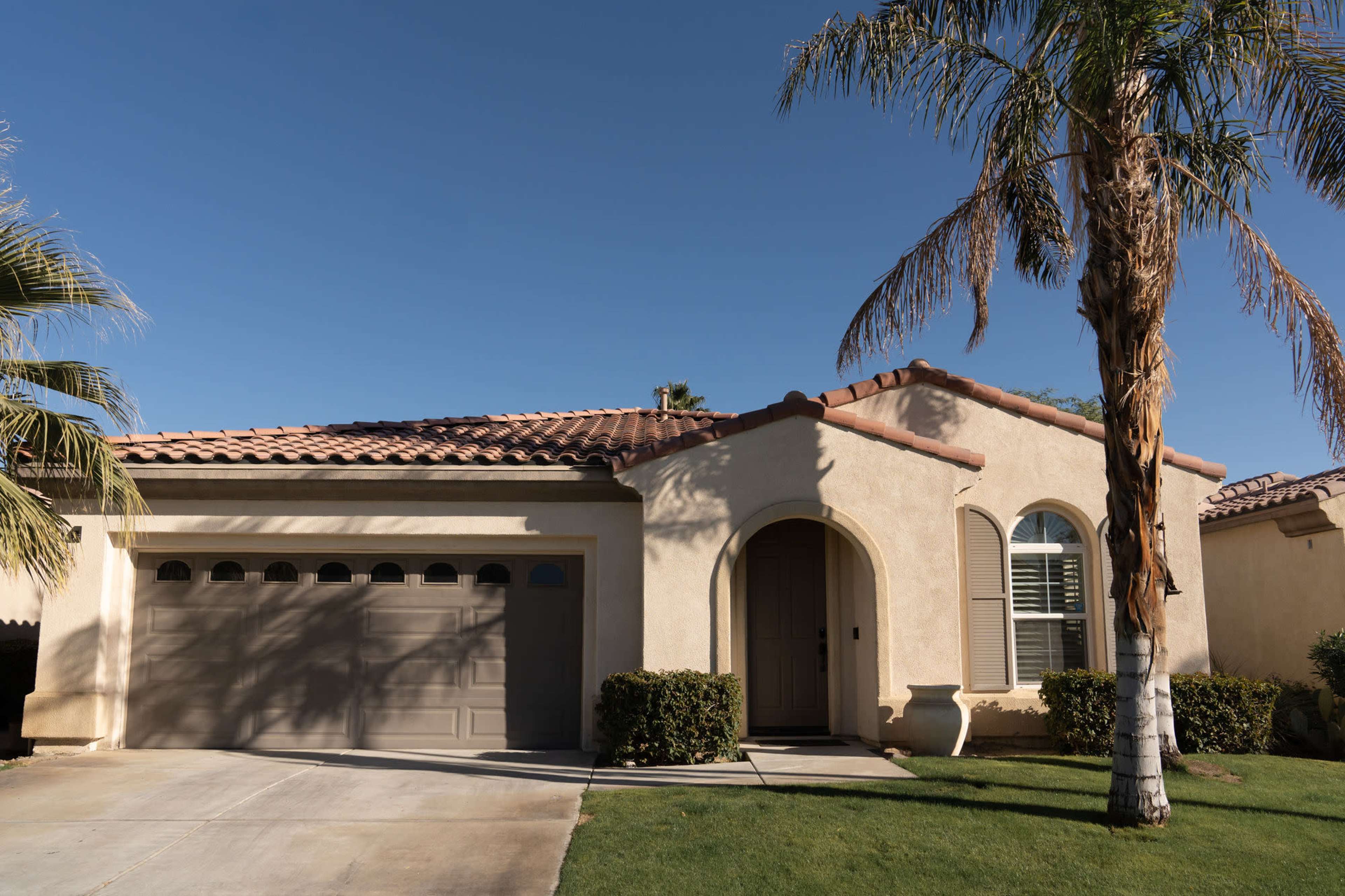 A single-story stucco house with a tile roof, a garage on the left, and palm trees in the front yard.