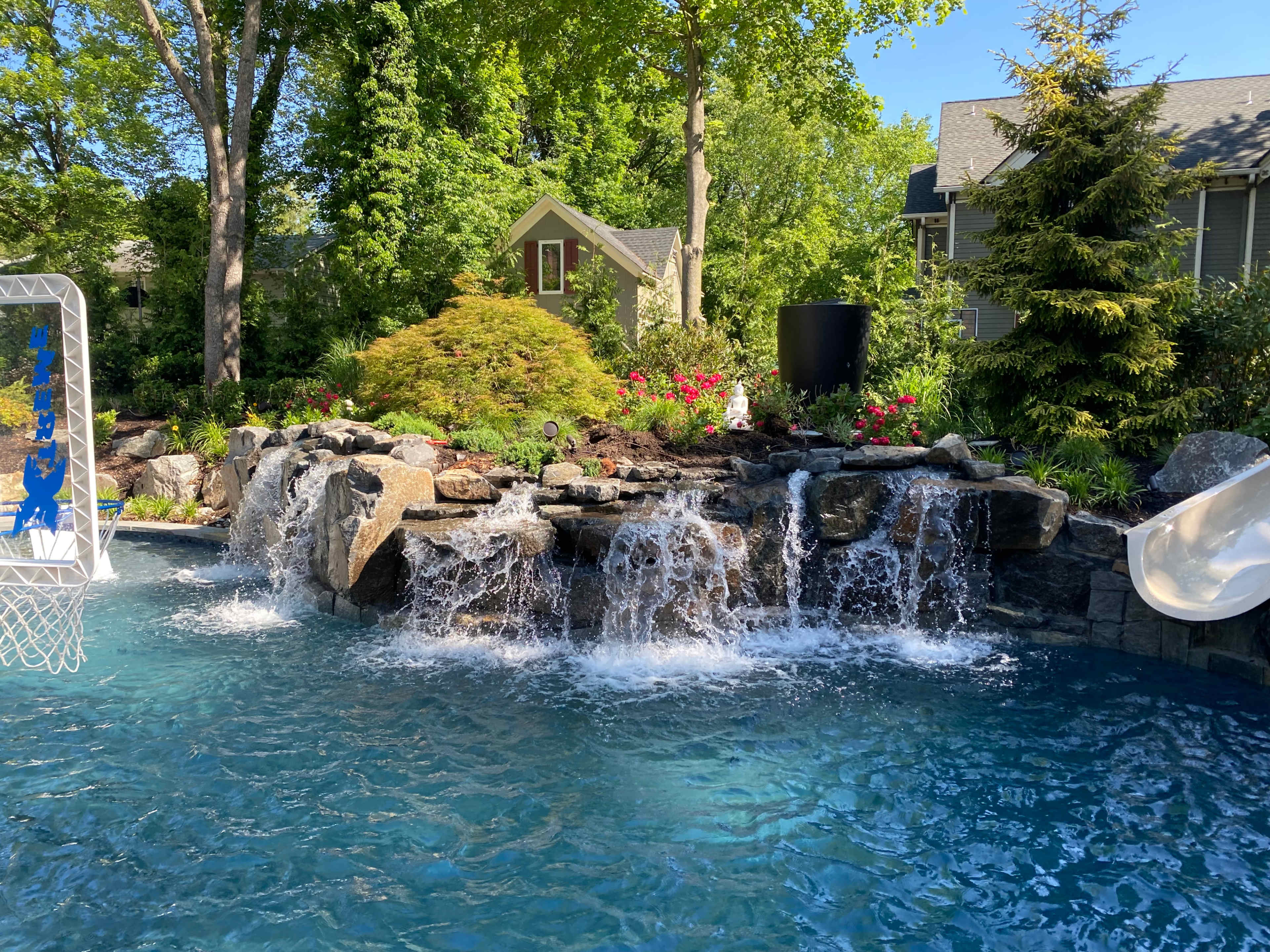 The scene features a backyard swimming pool with a rock waterfall, surrounded by lush greenery and colorful flowers.
