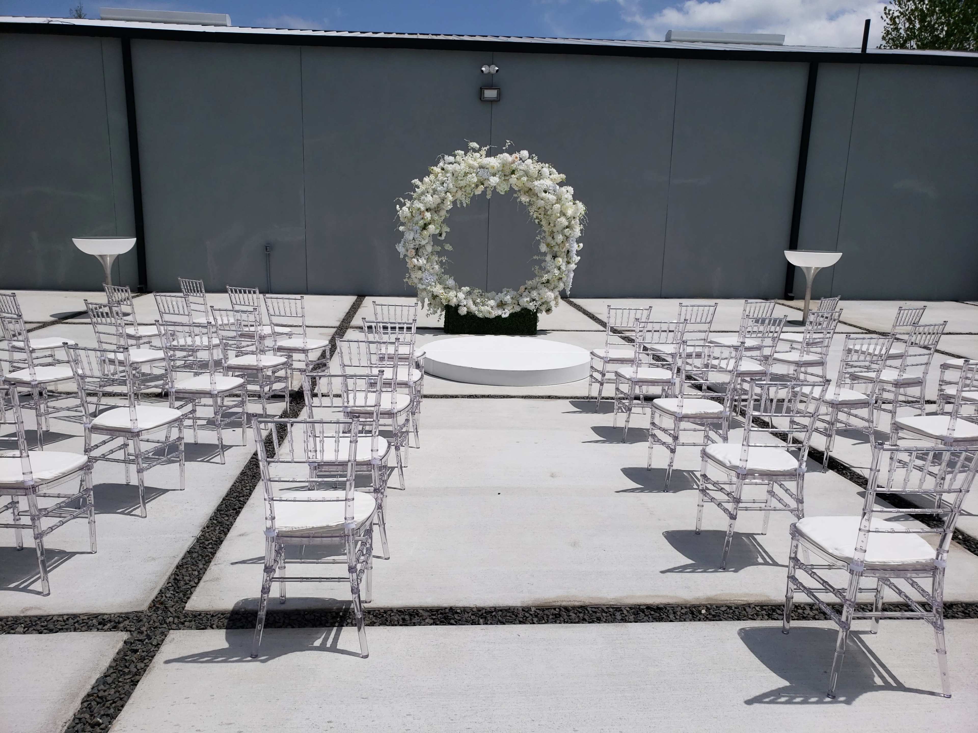 The image shows a wedding ceremony setup featuring white chairs arranged in a circular pattern around a floral arch on a raised platform.