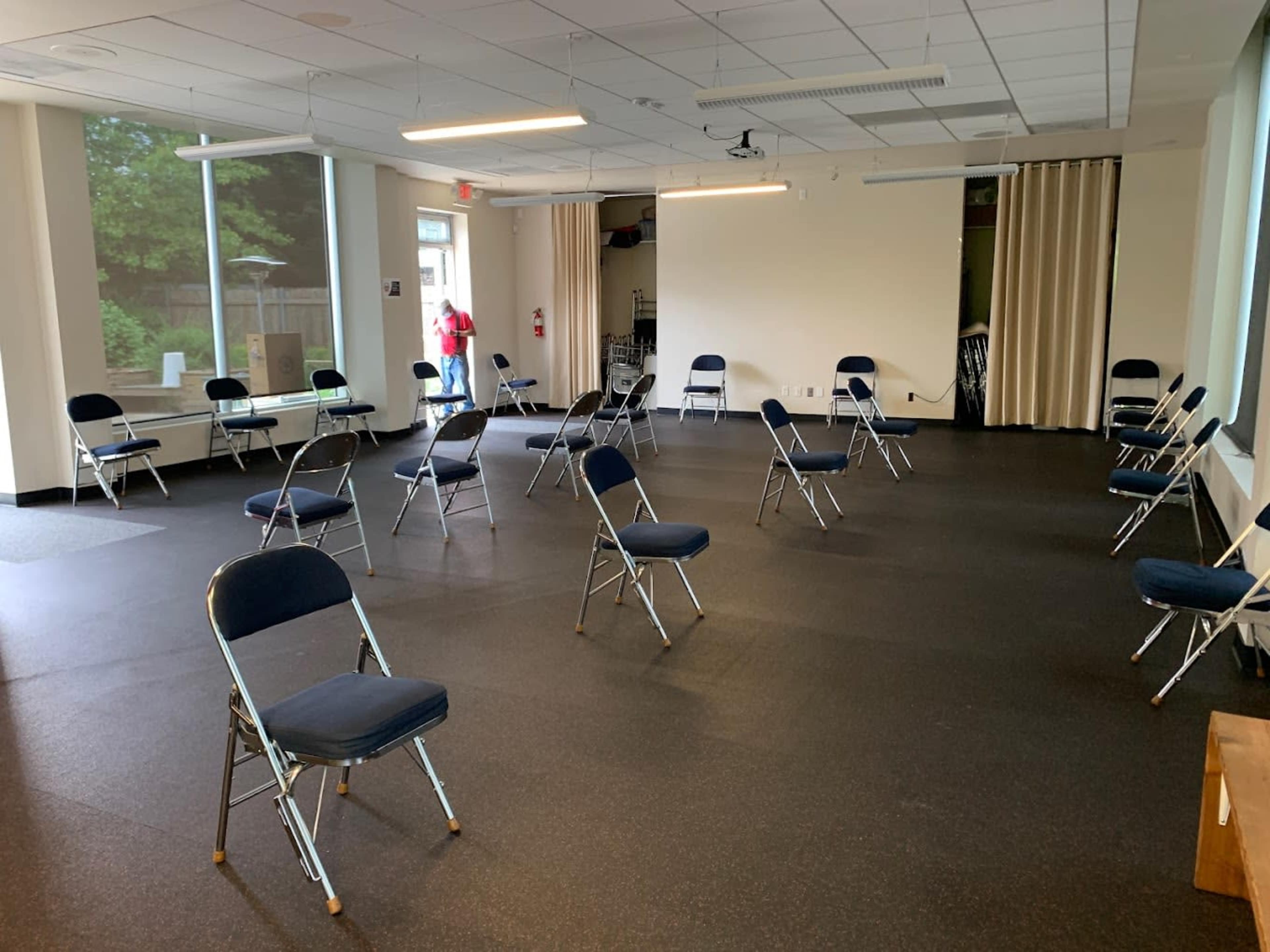 A large, empty room with blue folding chairs arranged in a spaced-out configuration on a black floor.