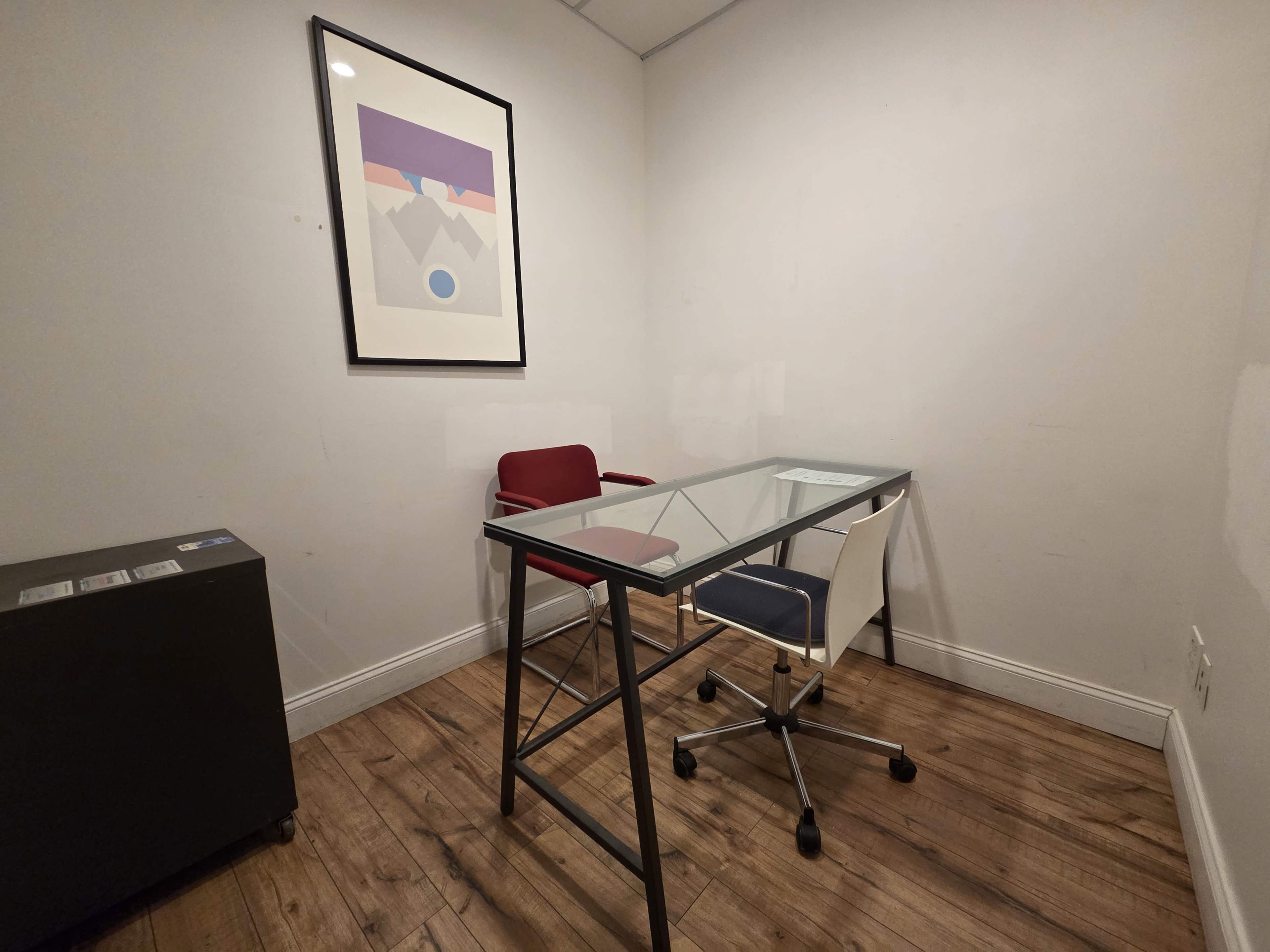 The image shows a small office space featuring a glass-topped desk, two chairs in red and blue, and a framed artwork on the wall.