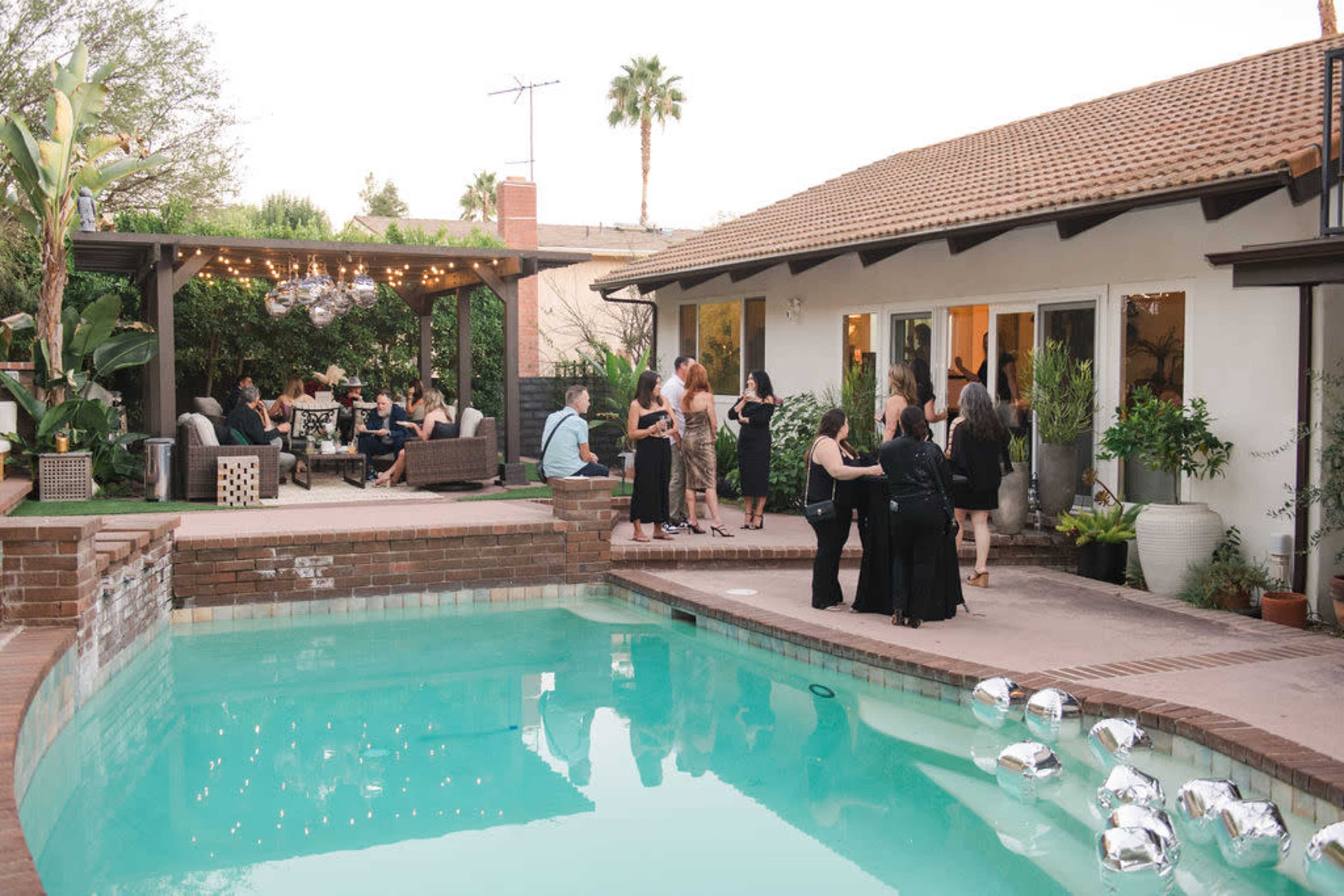 A group of people socialize by a poolside at a gathering in a backyard, with lounge furniture and decorative lighting visible.