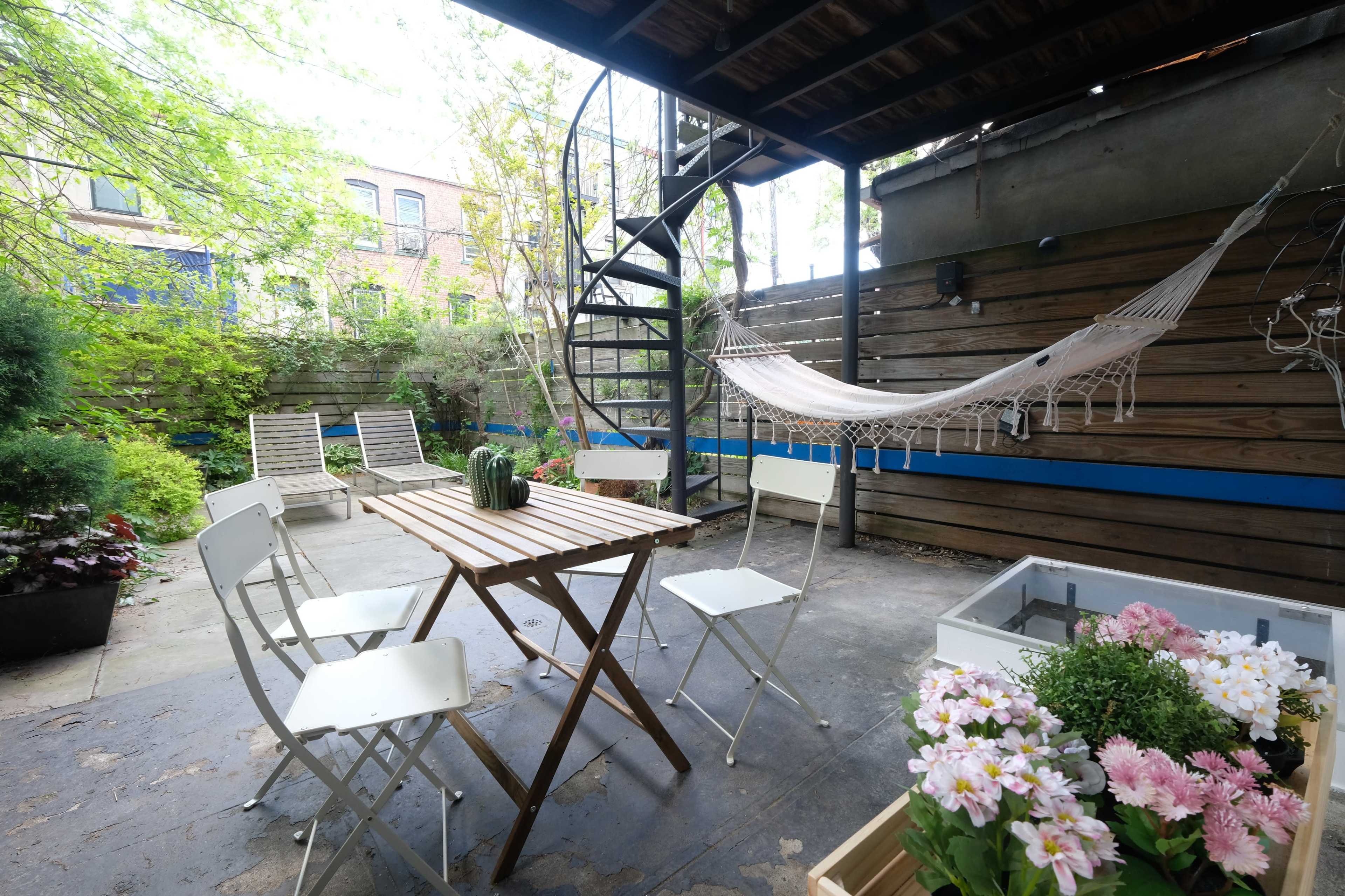 The image shows a small outdoor patio area with a spiral staircase, a wooden table surrounded by white chairs, a hammock, and potted flowers.
