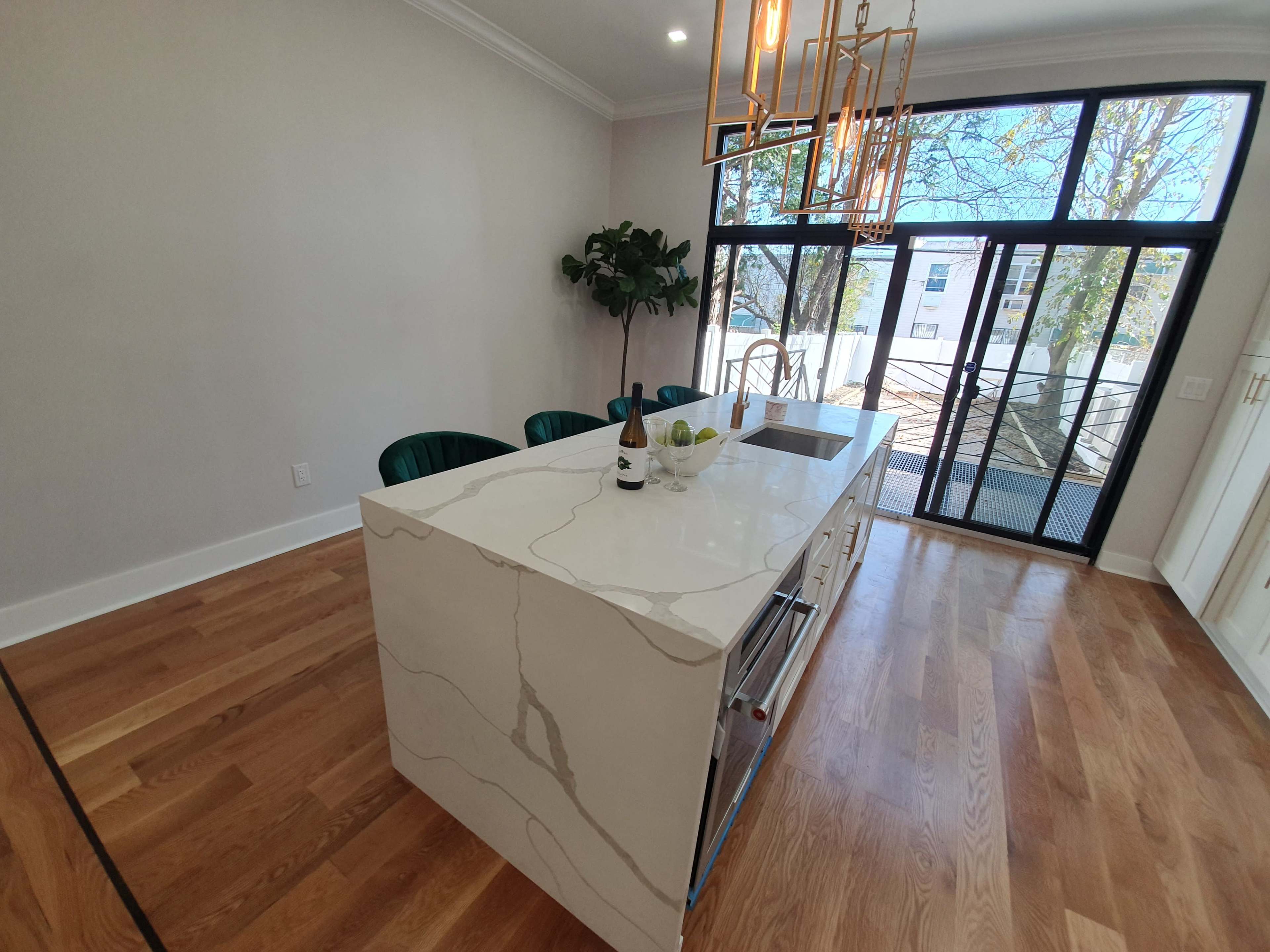 A modern kitchen features a large marble island with a wine bottle and fruit bowl, surrounded by green chairs, with large windows opening to a patio.