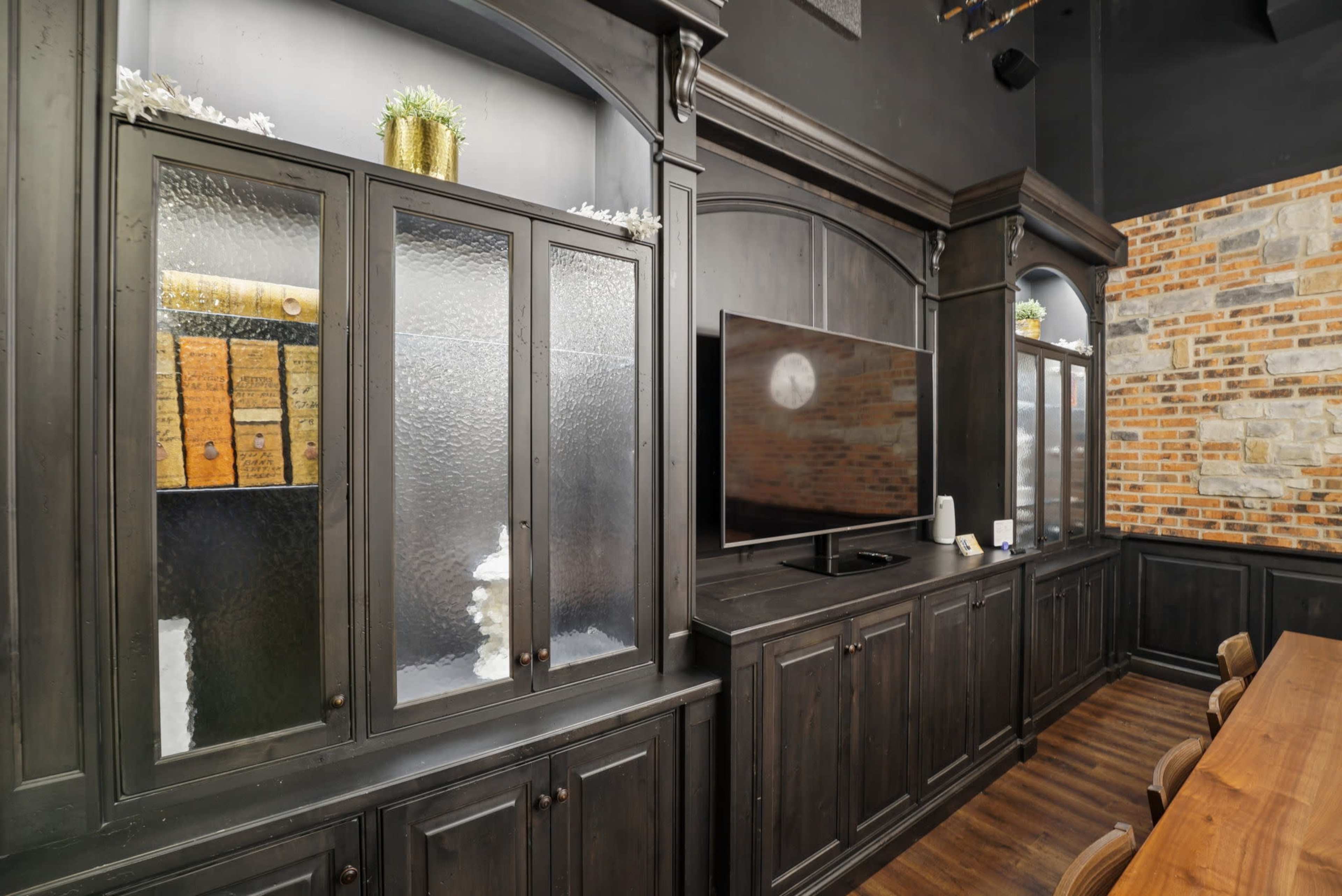 The image shows a dark wood cabinet with glass doors, displaying neatly arranged items, next to a large television mounted on the wall in a modern interior with stone accents.