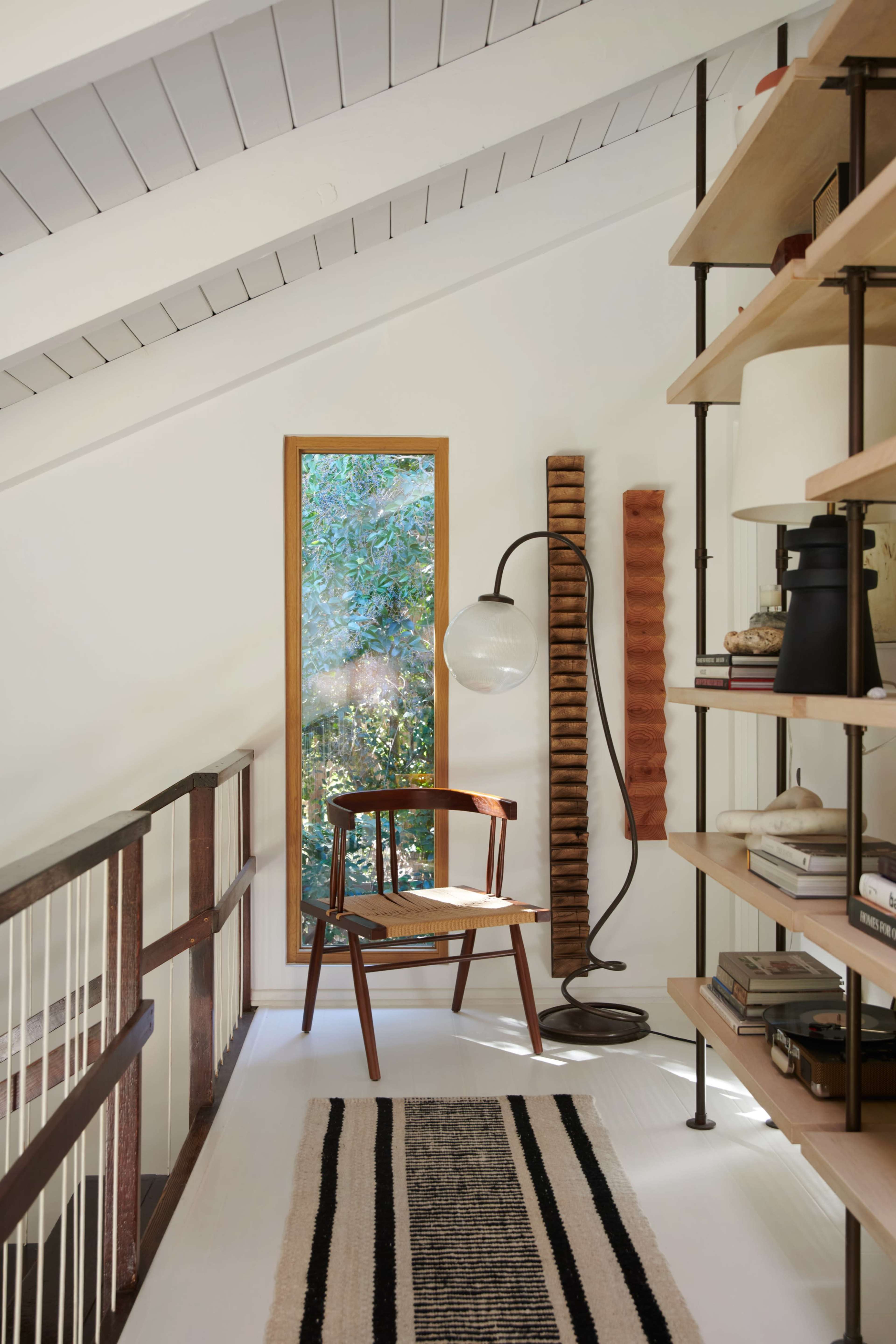 A wooden chair is placed beside a modern floor lamp in a bright hallway, with a large window offering a view of greenery and a bookshelf filled with various items.
