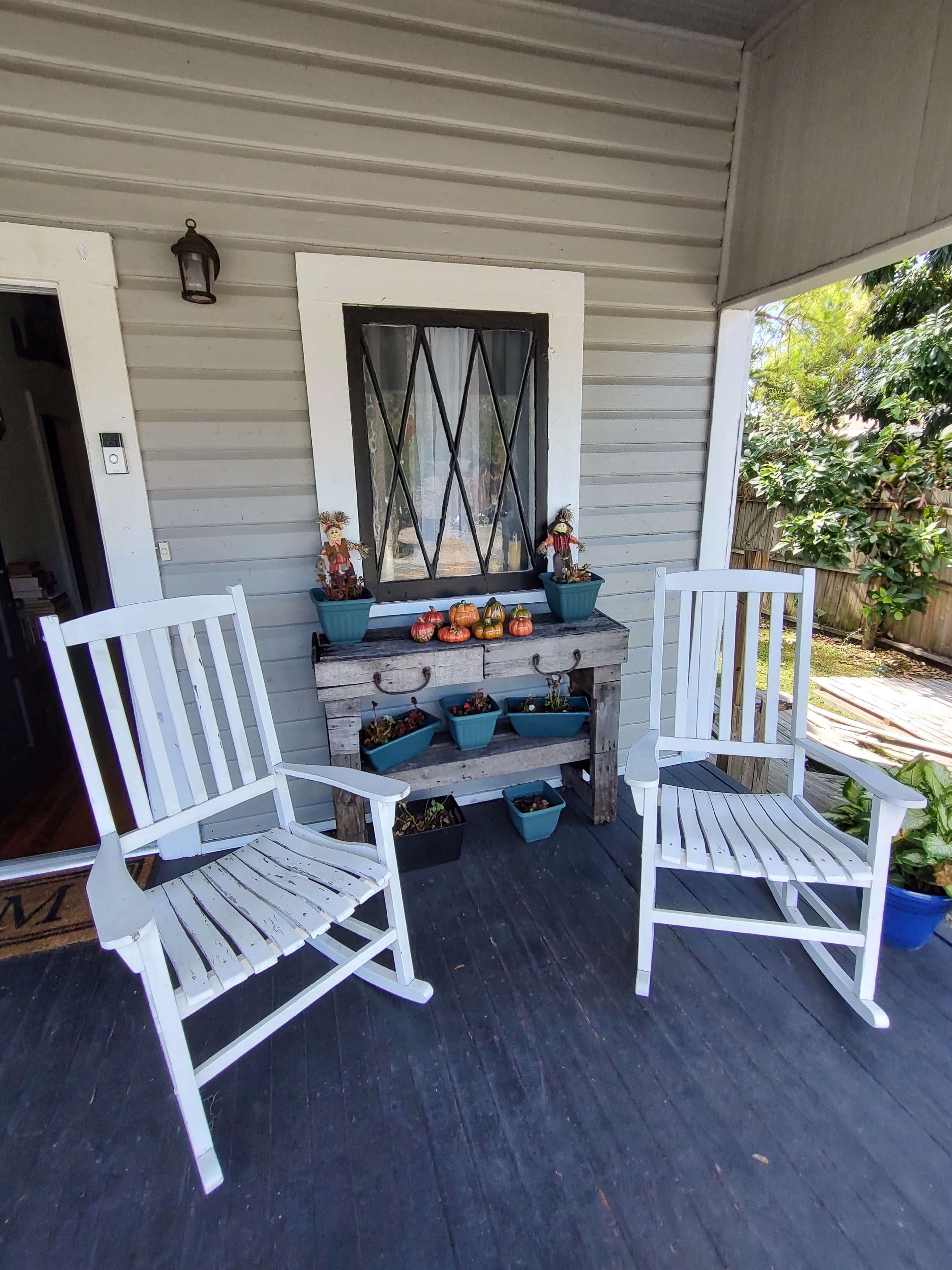 Two white rocking chairs sit on a wooden porch in front of a table adorned with potted plants and decorative items.