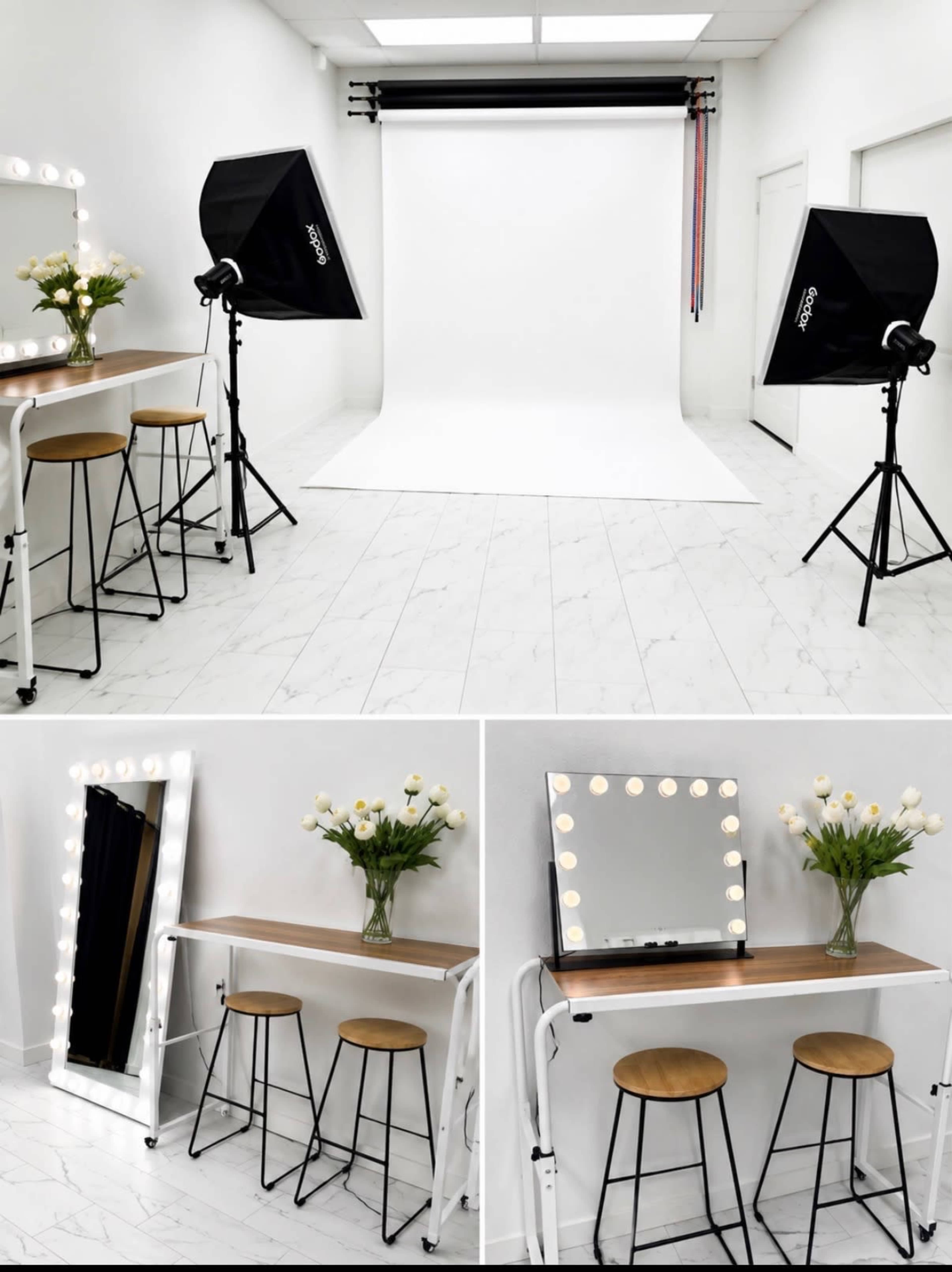 The image shows a well-lit photo studio featuring a white backdrop, two light stands with softboxes, a narrow table with a mirror, and three bar stools.