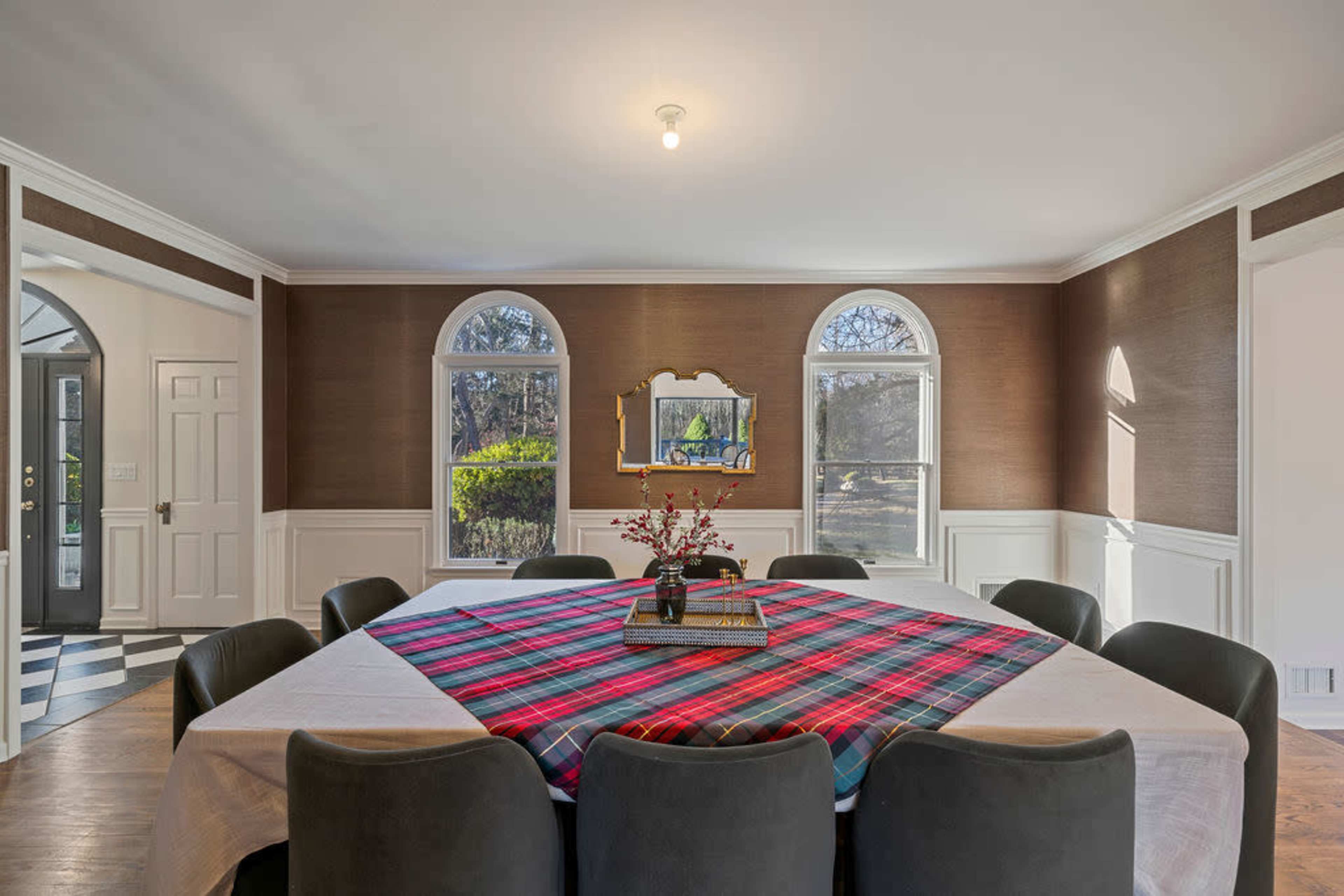 A spacious dining room features a large table covered with a plaid tablecloth, surrounded by dark chairs, and is illuminated by a central light fixture.
