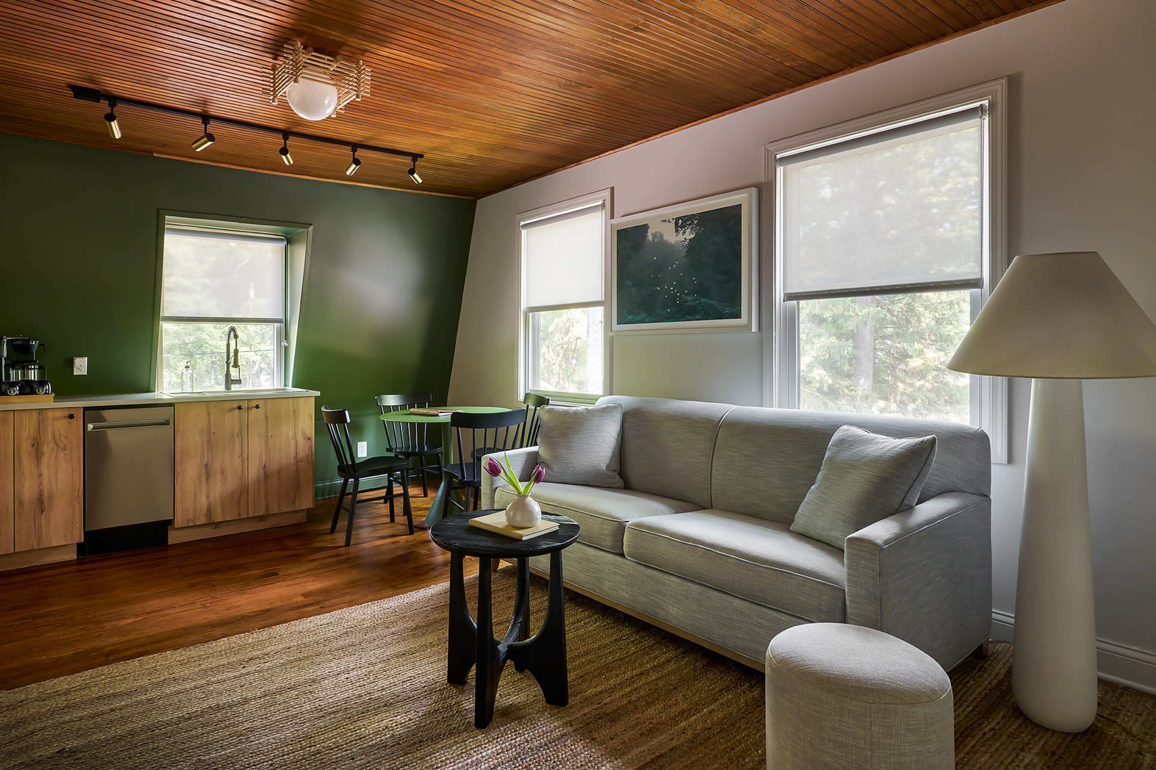 A cozy living area with a gray sofa, a small black side table with a vase of flowers, and a kitchenette featuring wooden cabinets in a green-painted room.