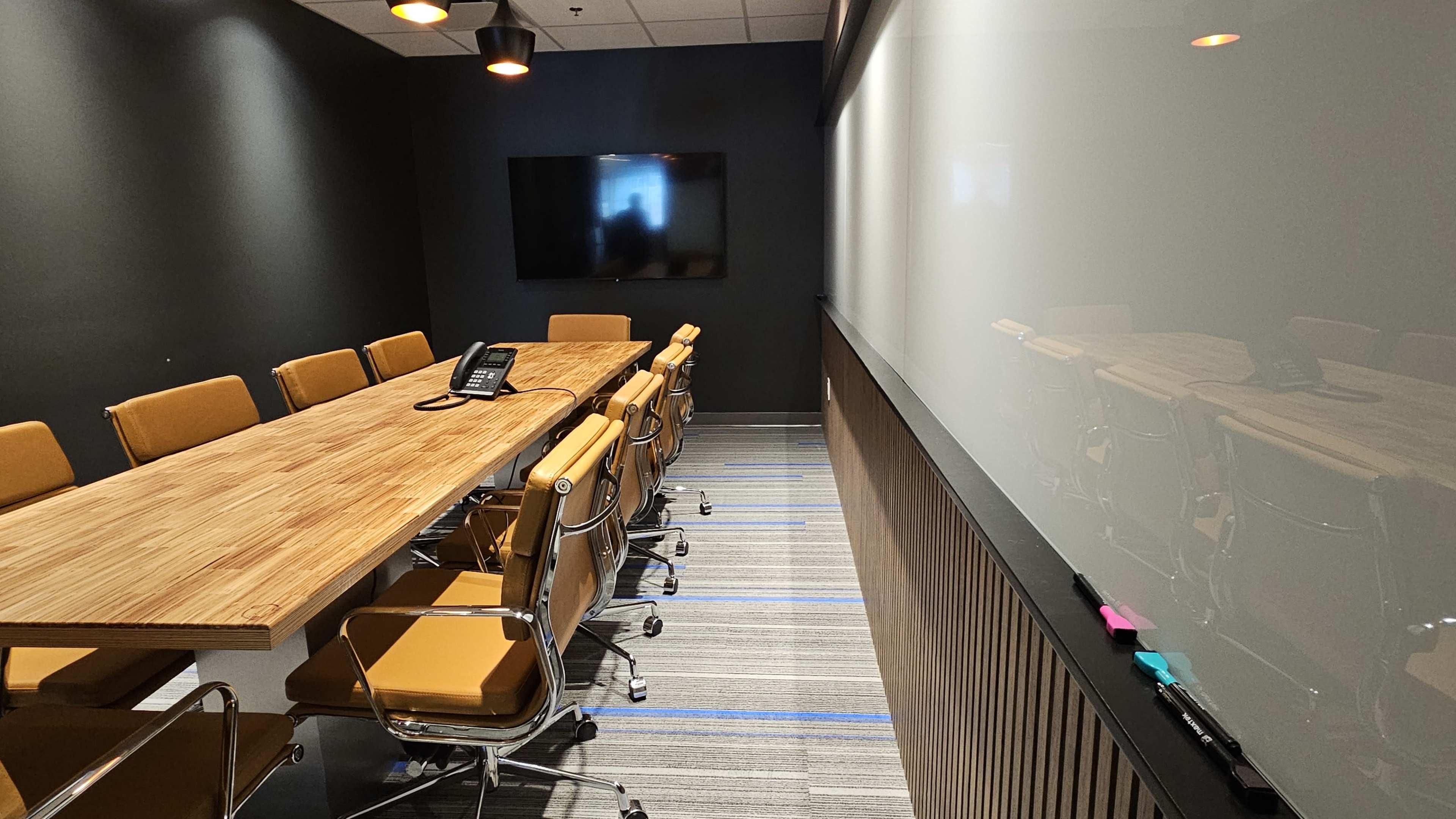 A conference room featuring a large wooden table surrounded by orange leather chairs, a wall-mounted television, and a whiteboard.