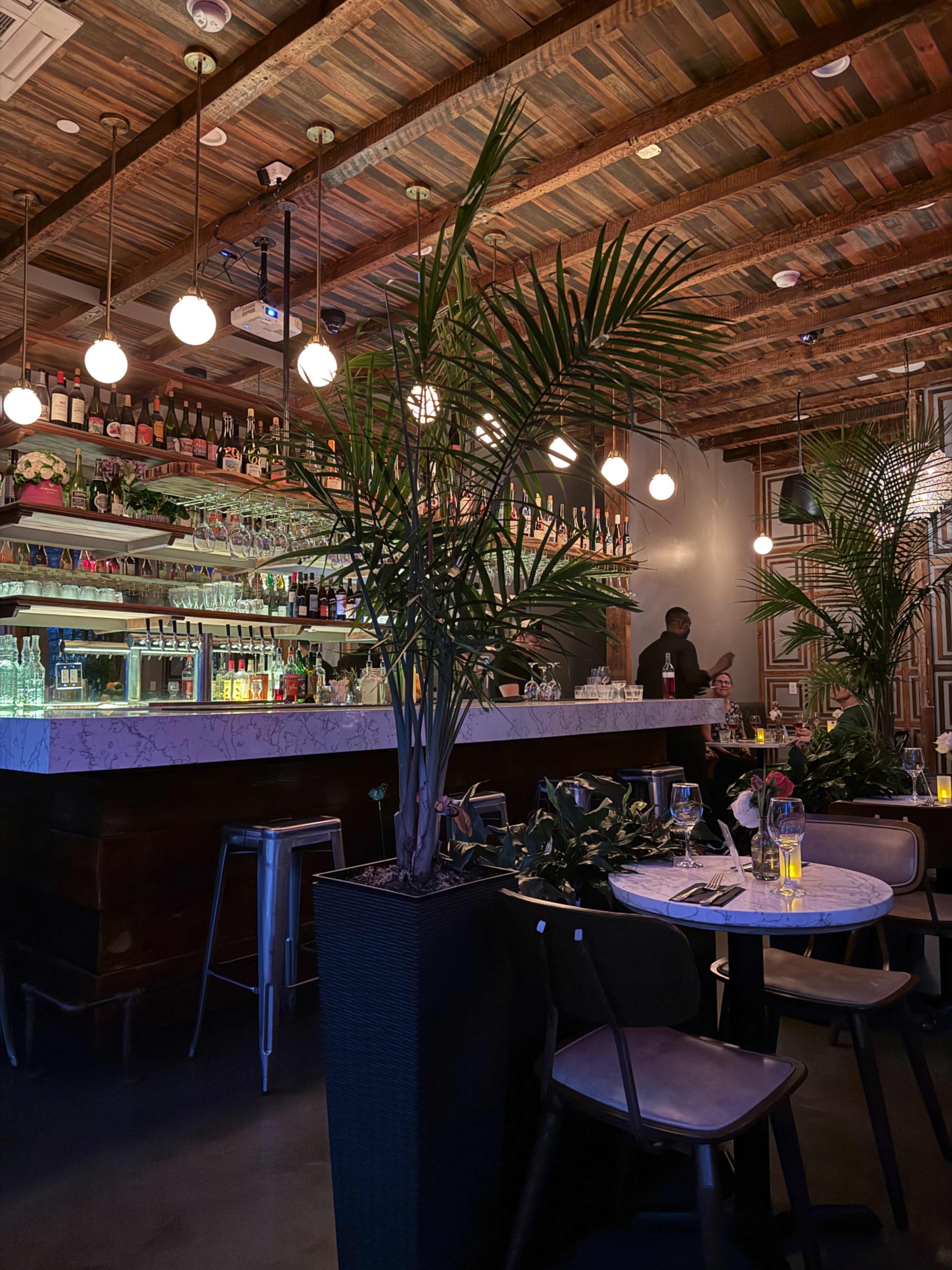 The image shows a modern restaurant interior featuring a bar with shelves of bottles, a marble countertop, and plants, with dim lighting and a few patrons seated at tables.