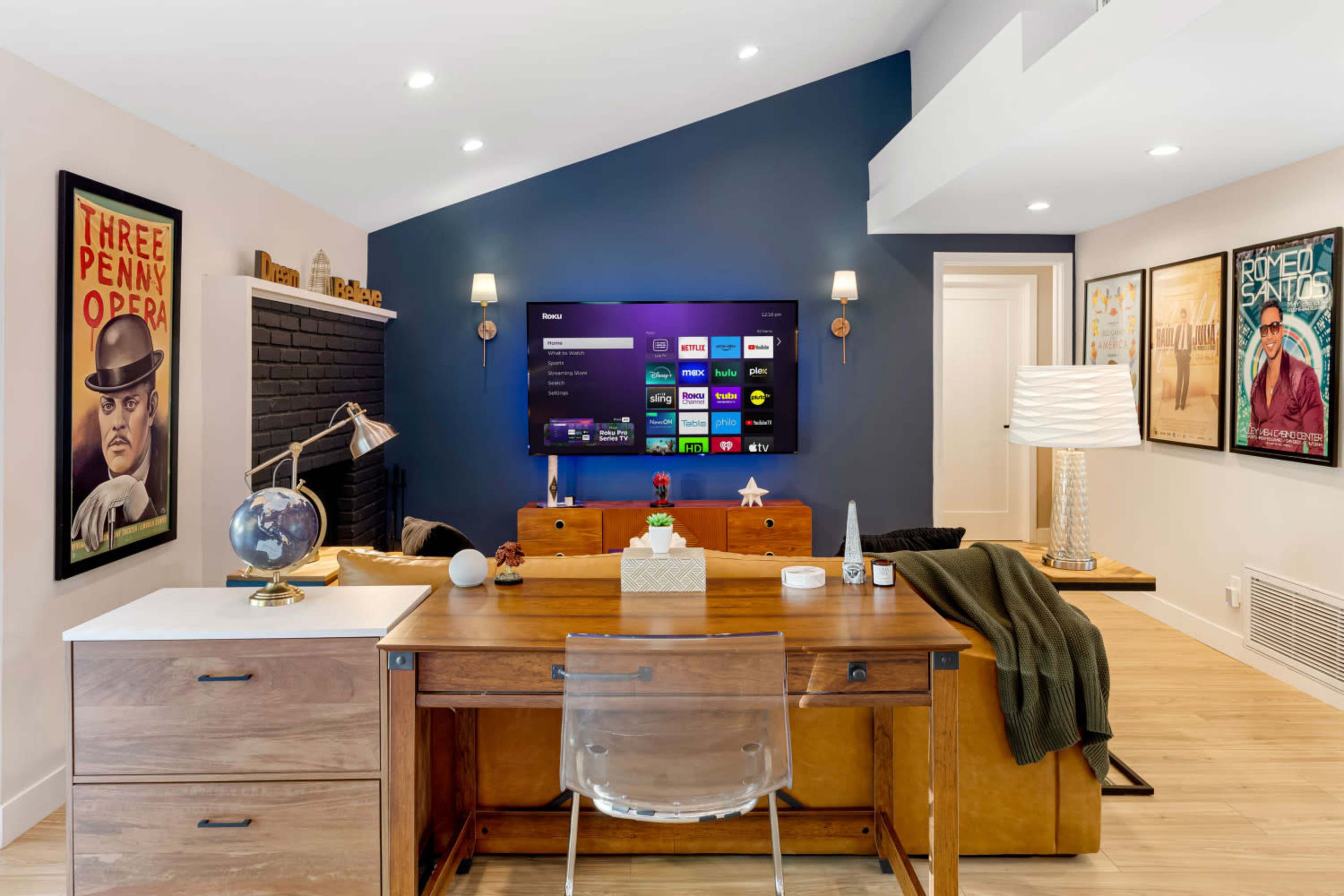 The image shows a modern home office with a wooden desk, a television mounted on a blue accent wall, and decorative artwork on the surrounding walls.