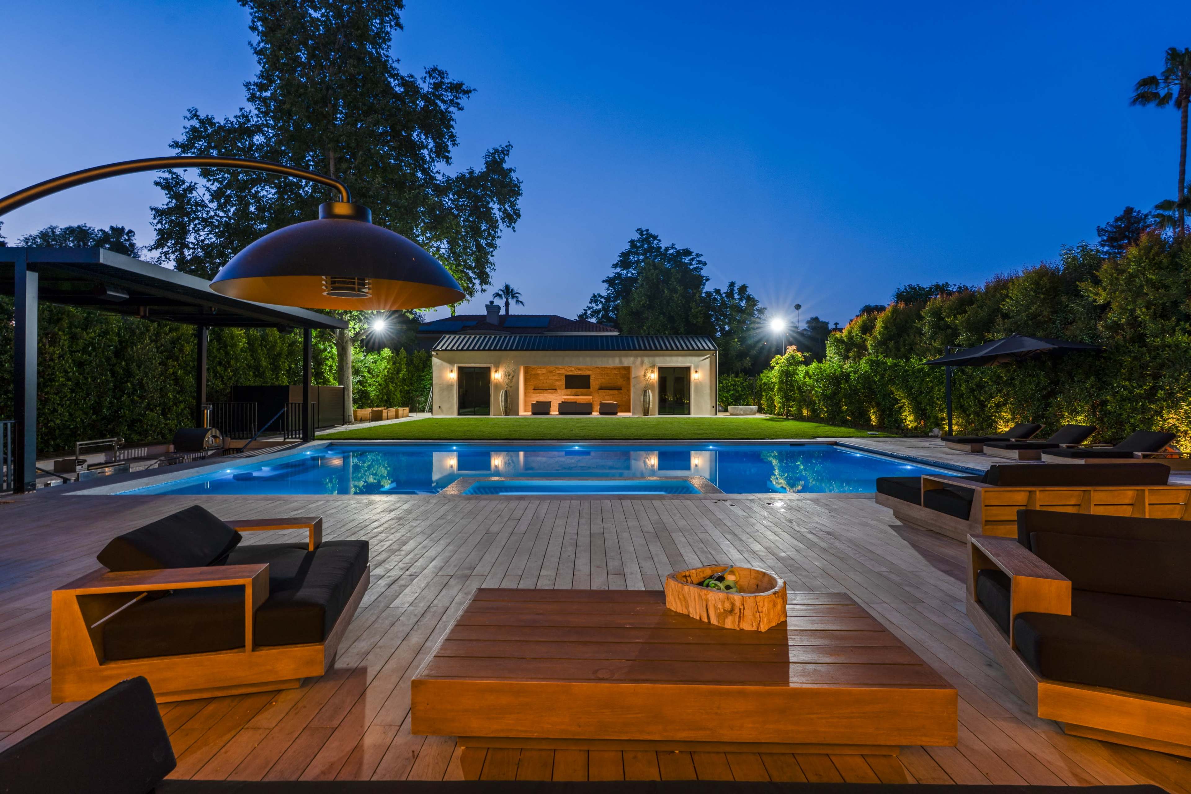 A modern backyard features a large swimming pool surrounded by wooden decking and lounge furniture, with a well-lit patio area and a distant view of a building.