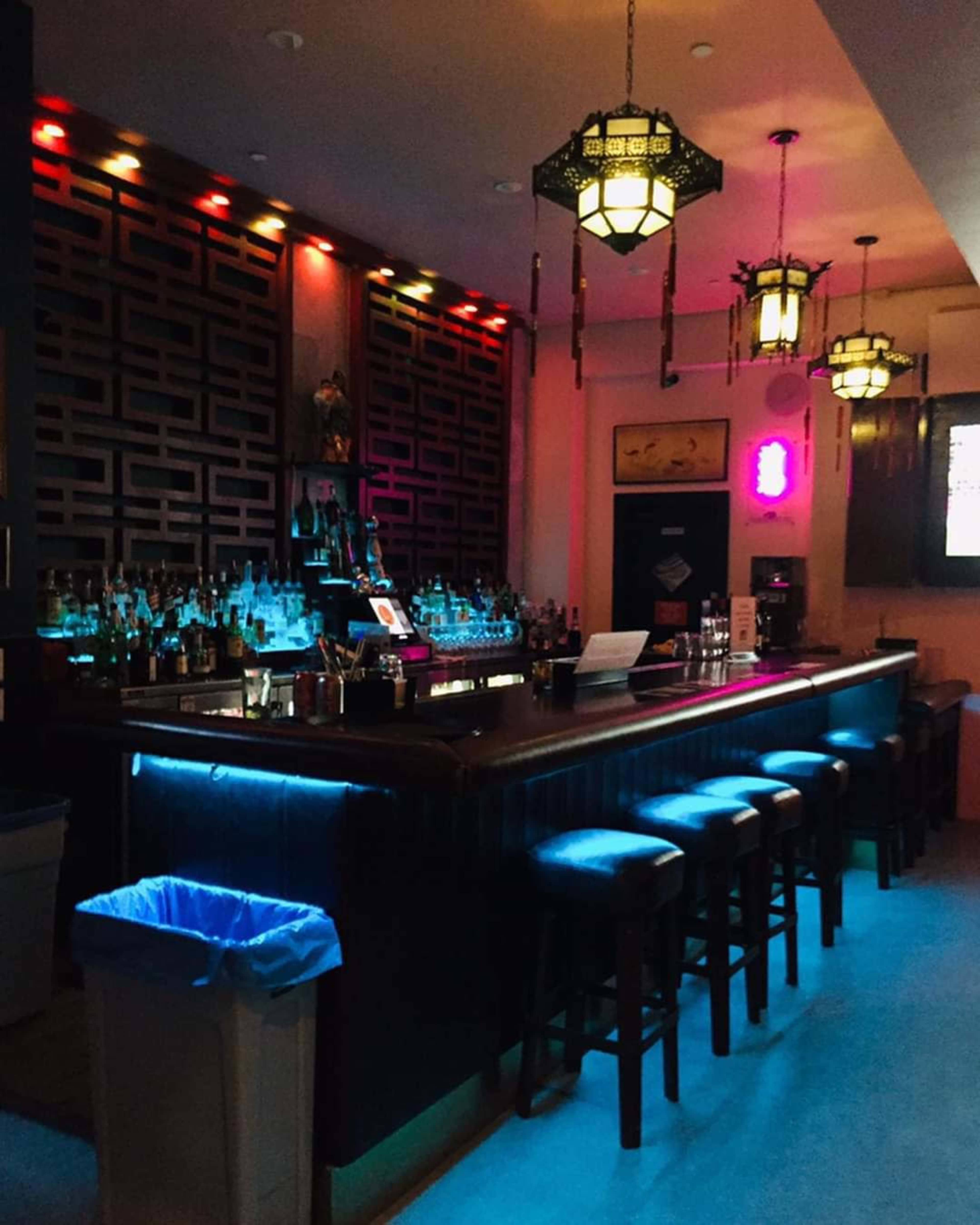The image shows a dimly lit bar with a long counter, illuminated by colorful lights and decorative lanterns.
