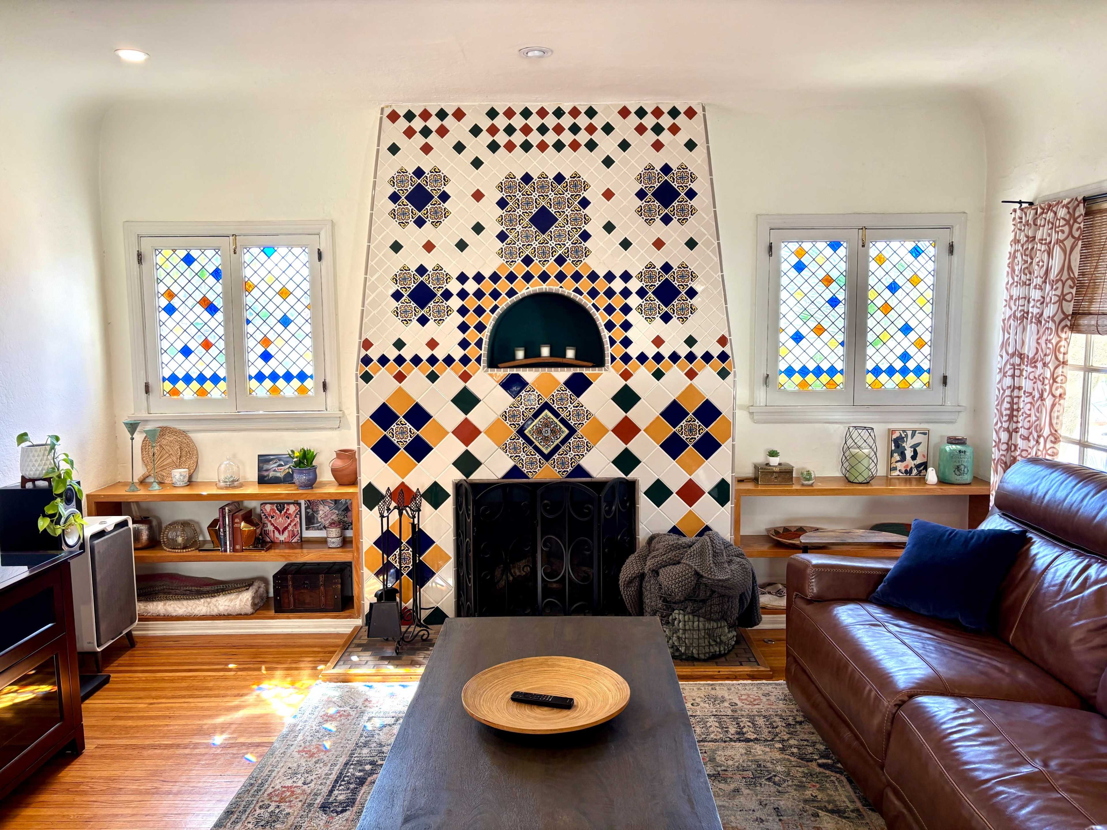 The image shows a living room featuring a decorative tiled fireplace, colorful stained glass windows, a wooden coffee table, and a brown leather sofa.