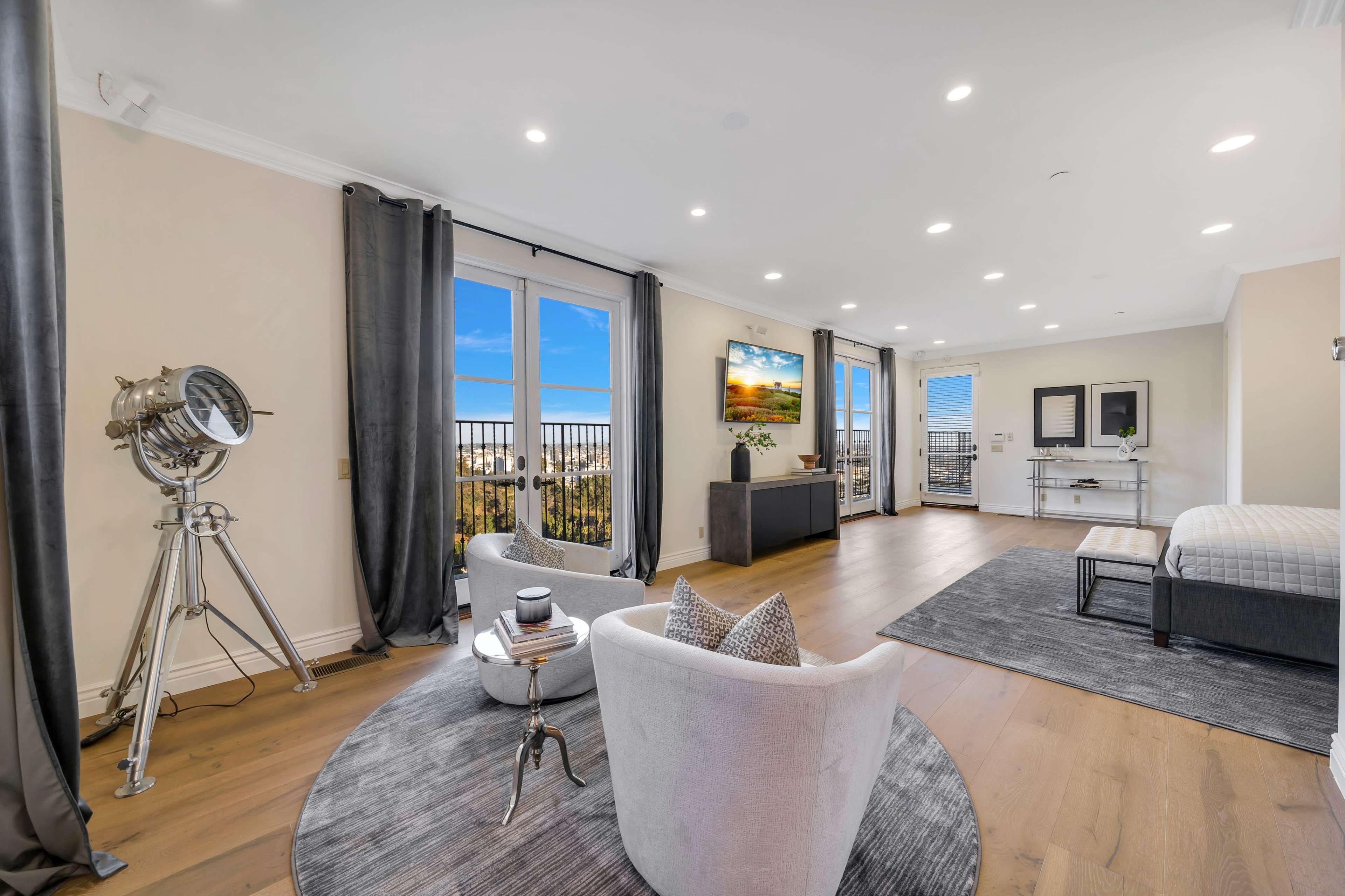 The image shows a modern living room with two armchairs, a large area rug, and a flat-screen TV mounted on a wall, featuring large windows that offer a view of the outdoors.