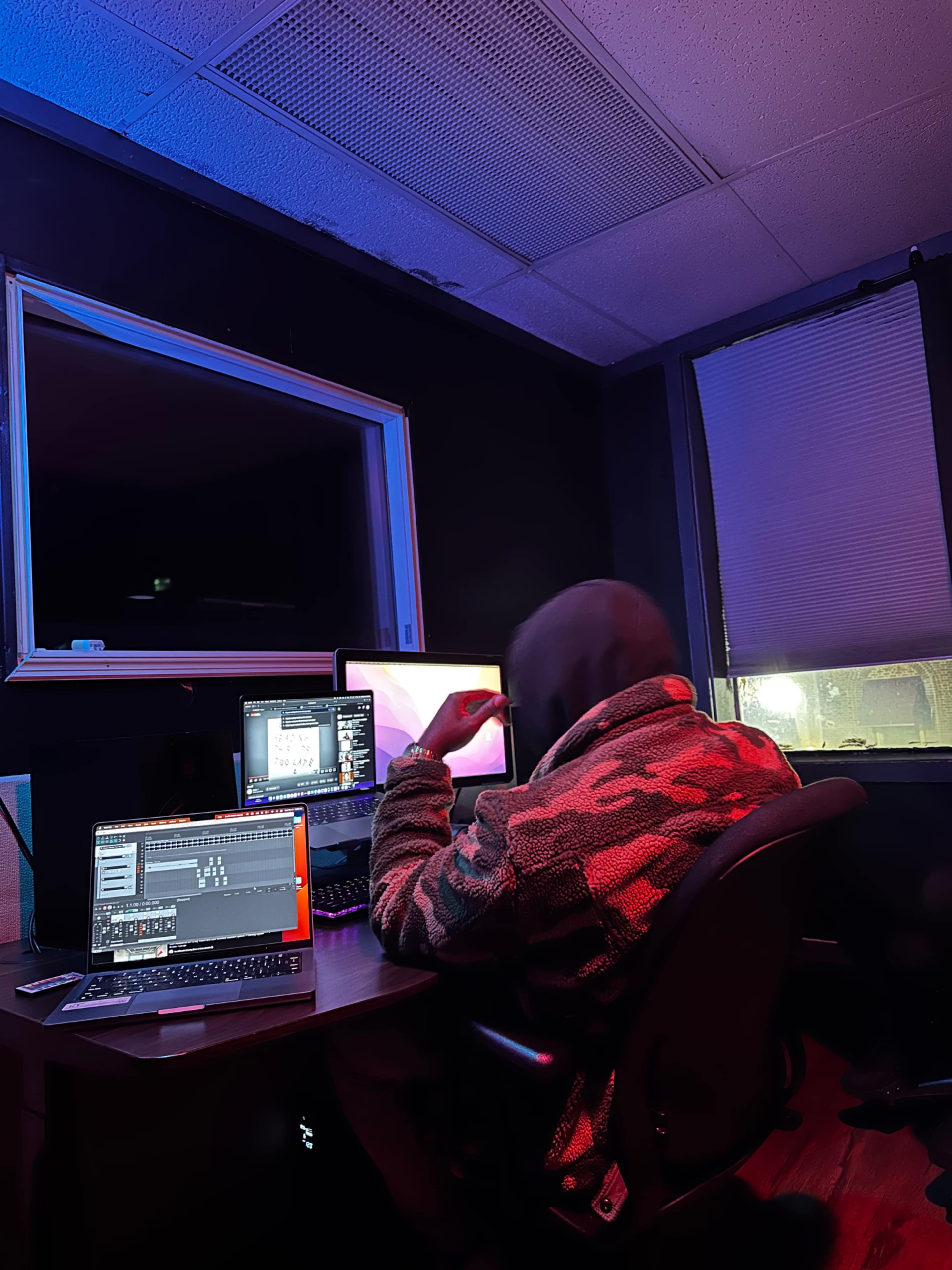A person in a hooded jacket sits at a desk in a dimly lit room, working on multiple computer screens.
