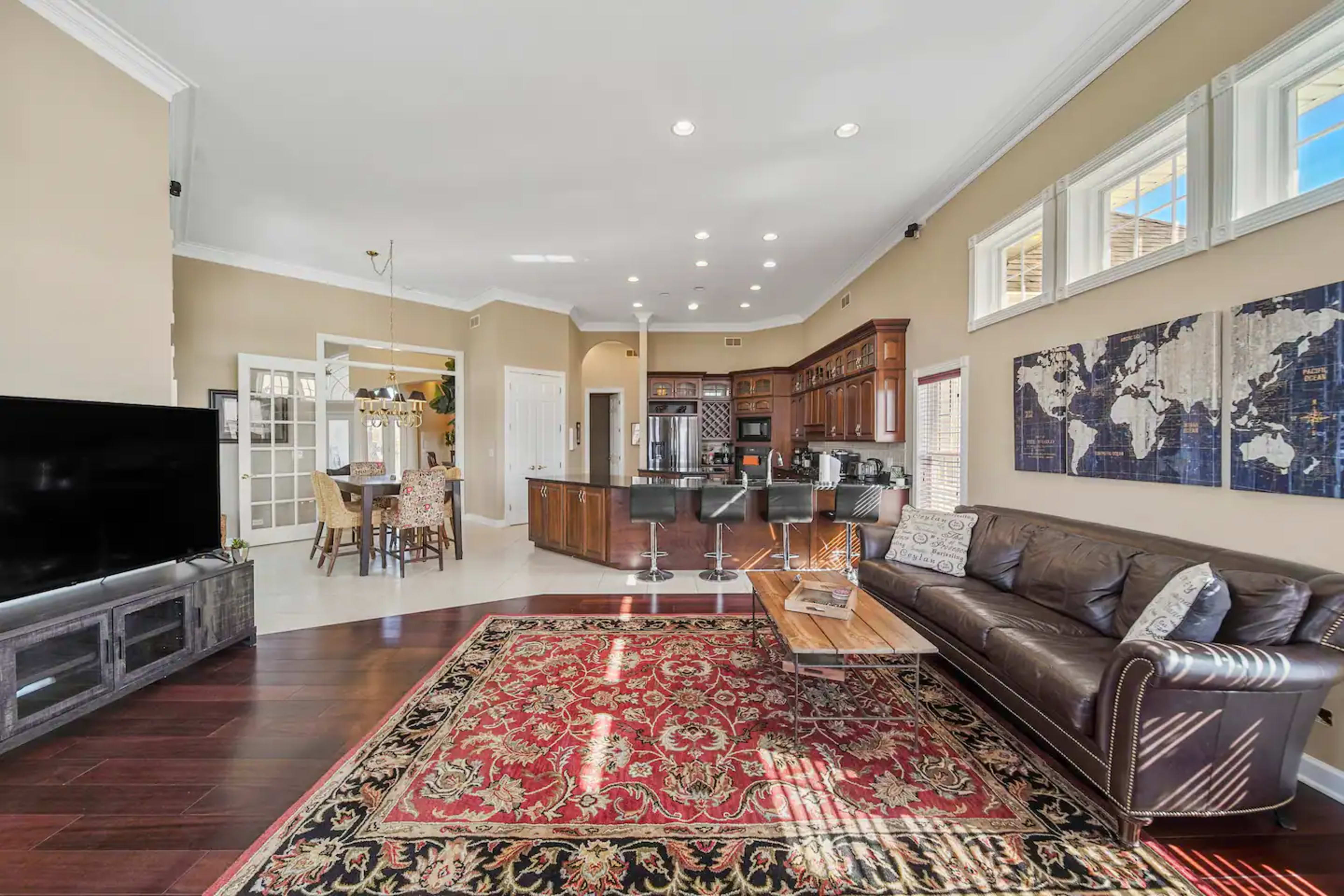 The spacious living area features a leather sofa, a large television, and an open kitchen with dark wood cabinetry, along with a dining area in the background.